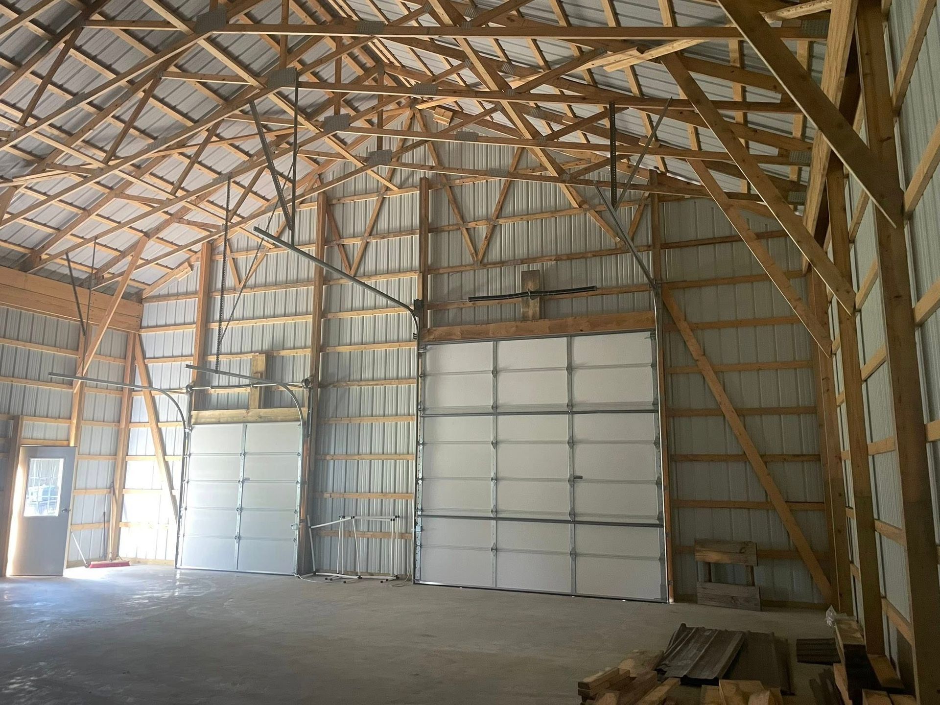 Interior of a large workshop with metal walls, wooden trusses, a concrete floor, and two white garage-style doors.