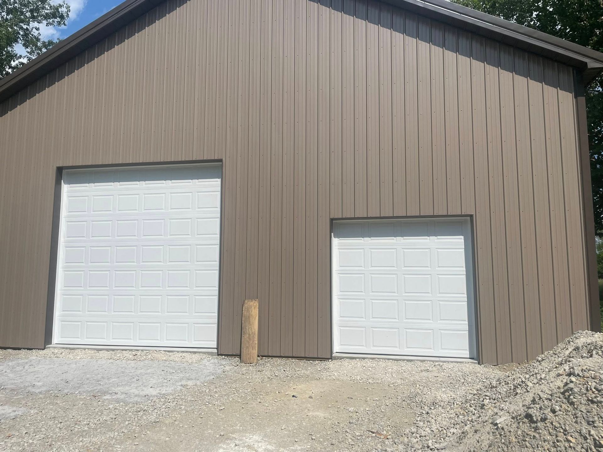 A brown metal building exterior features two white raised-panel garage doors of different sizes on a gravel lot.