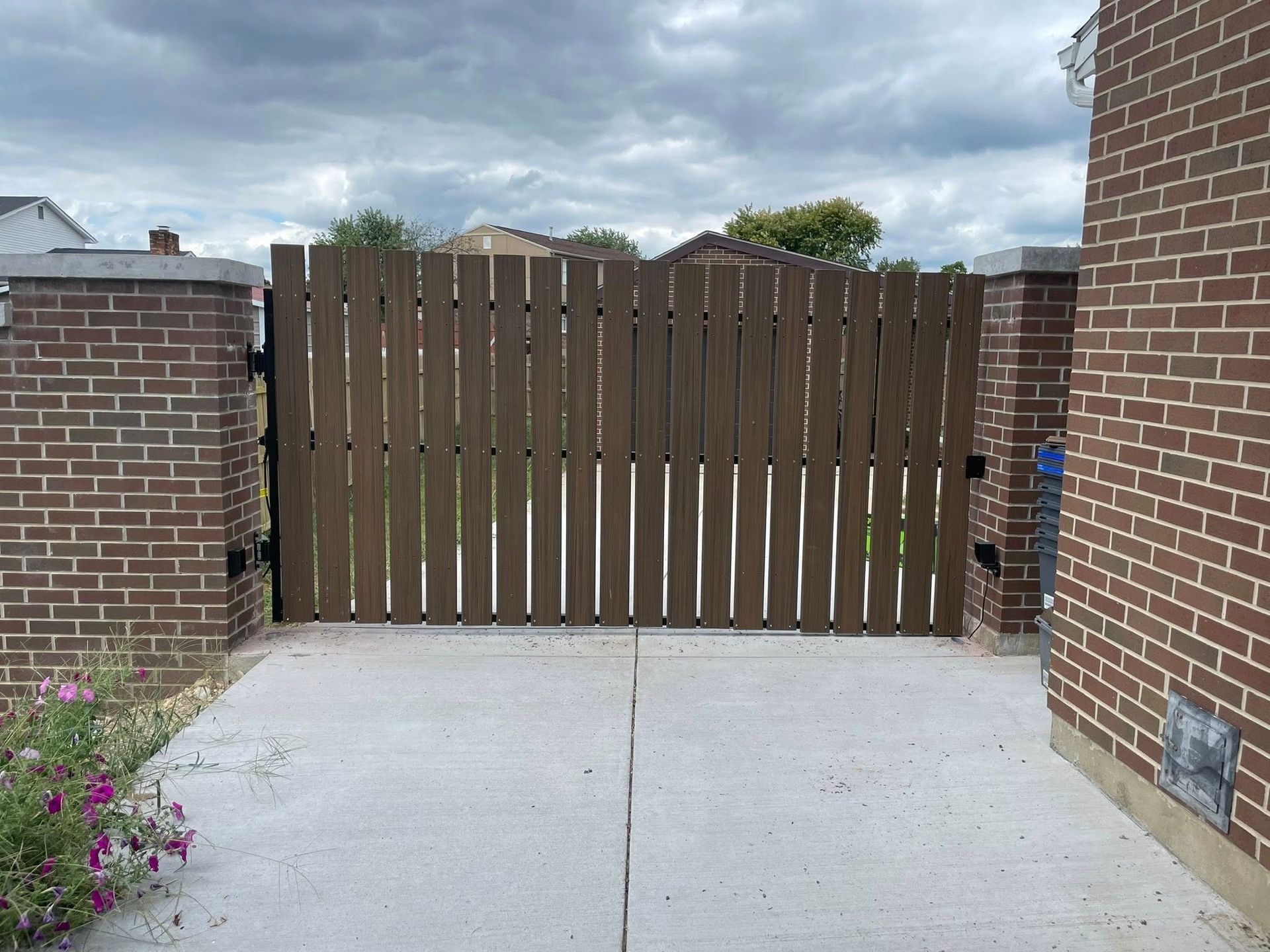 A wooden privacy gate with vertical slats sits between two brick pillars in a residential driveway under a cloudy sky.