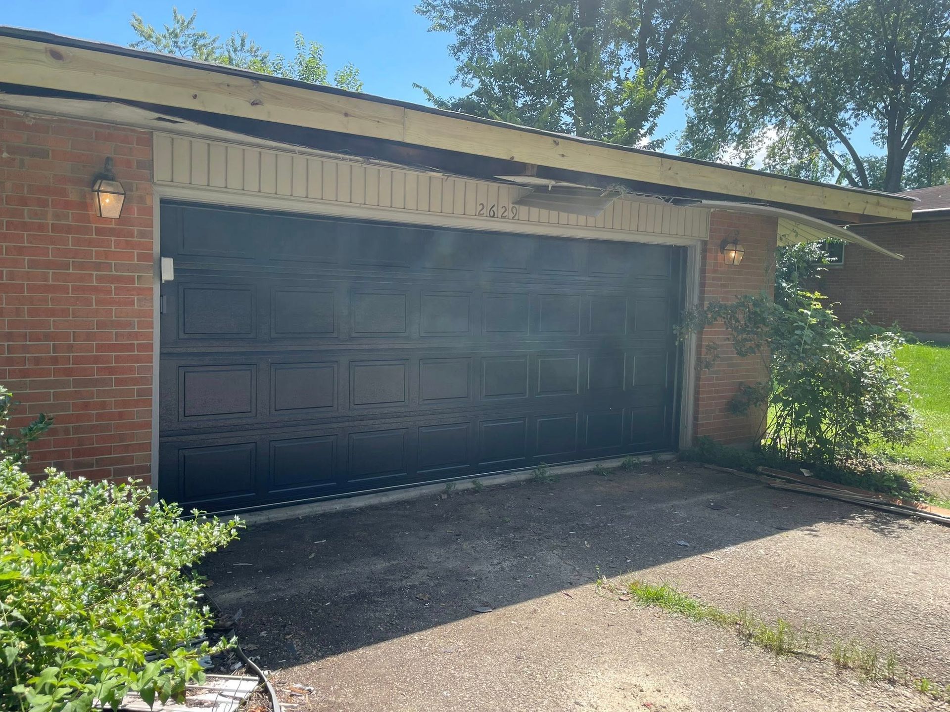 A black garage door on a brick building with a partially damaged wooden overhang above it under a bright blue sky.