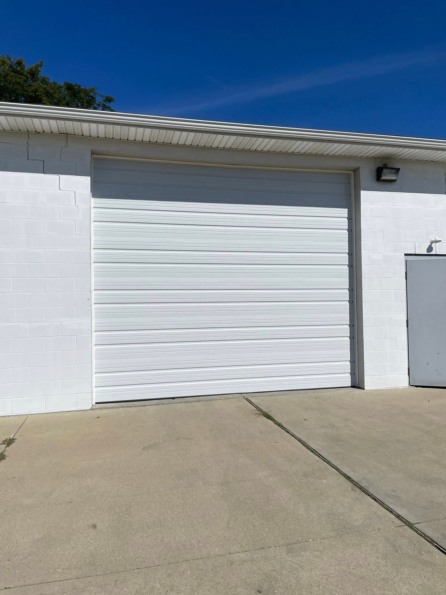 A large white roll-up garage door on a white concrete block building under a bright blue sky.