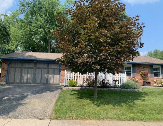A one-story brick house with a two-car garage, a front lawn, and a large reddish-leaf tree behind a white picket fence.