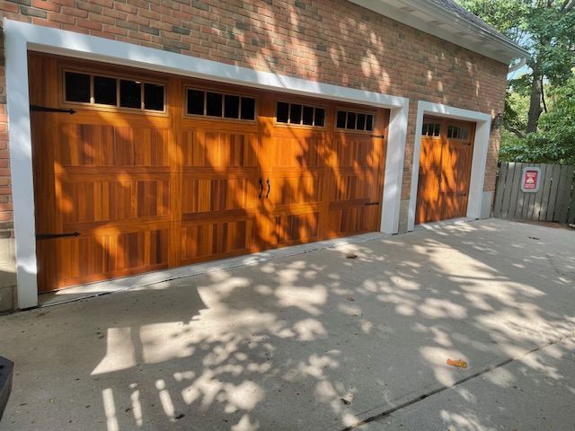 A detached brick garage with two wood-tone carriage-style doors under partial tree shade on a concrete driveway.