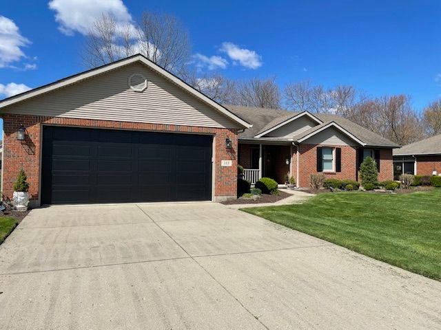 A one-story suburban home with tan siding, red brick accents, a black two-car garage, and a concrete driveway.