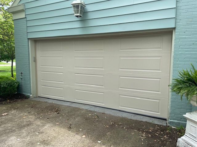 A beige, three-panel garage door installed in a light blue brick wall, set above a concrete driveway.