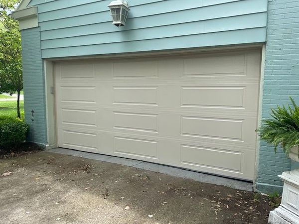 A beige, three-panel garage door installed in a light blue brick wall, set above a concrete driveway.