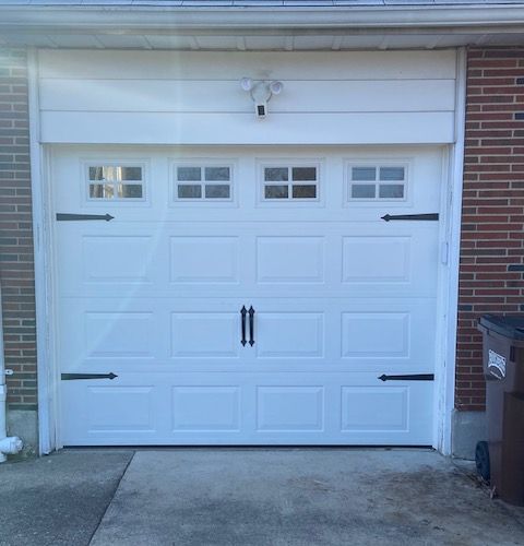 White garage door with four small windows, black decorative strap hinges, and a central handle, set in a brick frame.