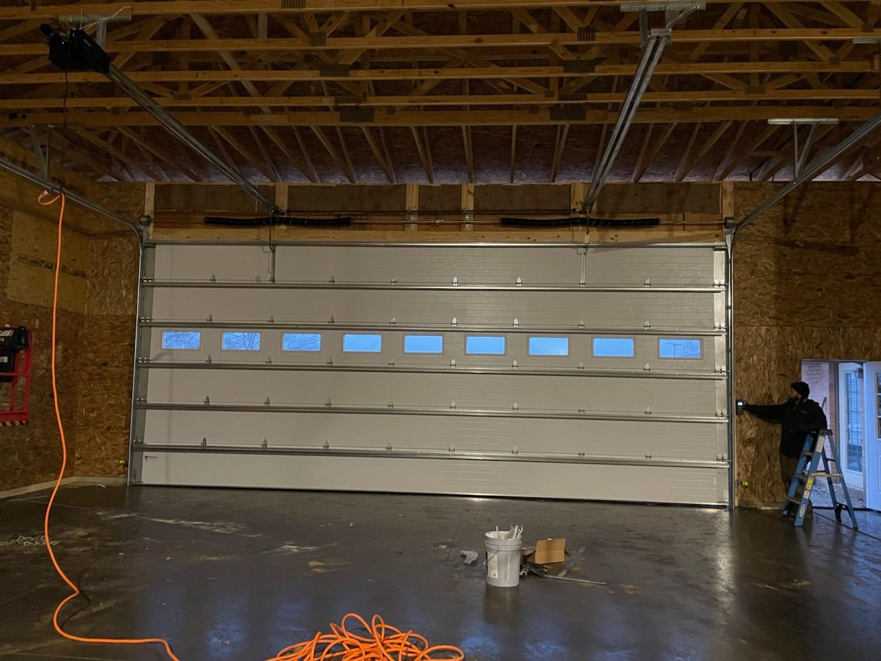 A person standing on a ladder next to a large, white, multi-paneled garage door inside an unfinished wooden workshop.