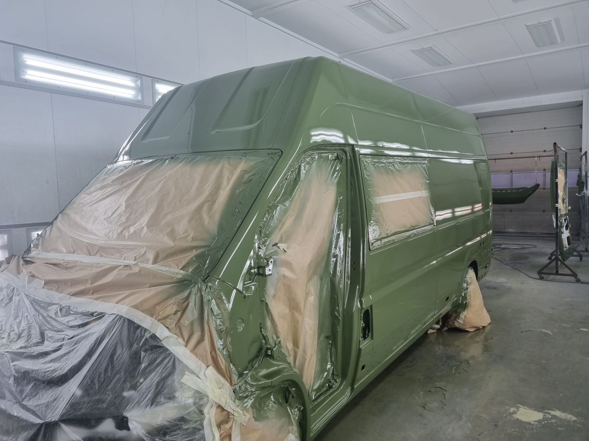 Een groene bestelwagen wordt in een spuitcabine gespoten; de ramen en de voorkant zijn met plastic afgedekt.
