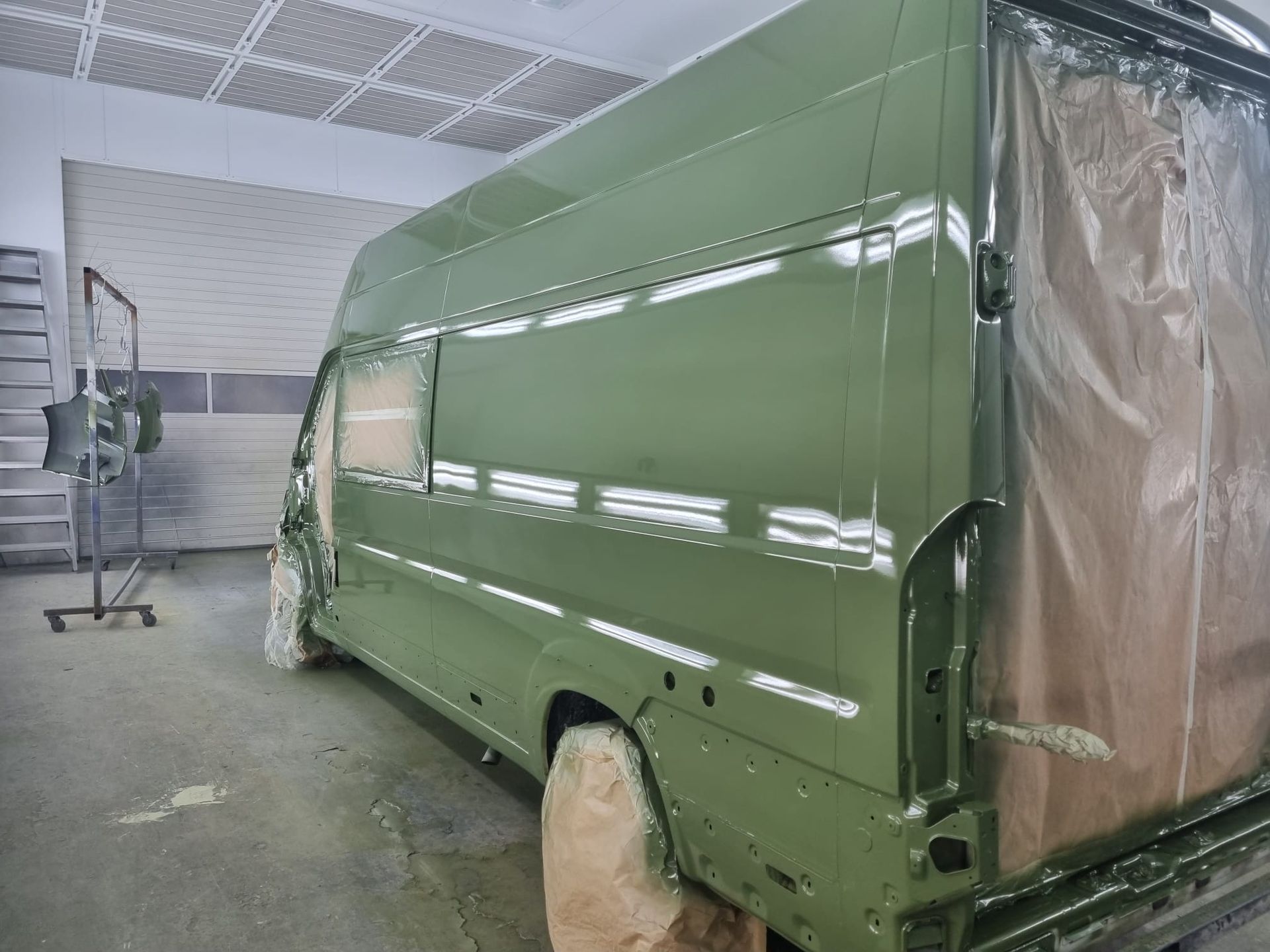 Een groene bestelwagen wordt in een werkplaats gespoten. Plastic folie beschermt bepaalde delen.