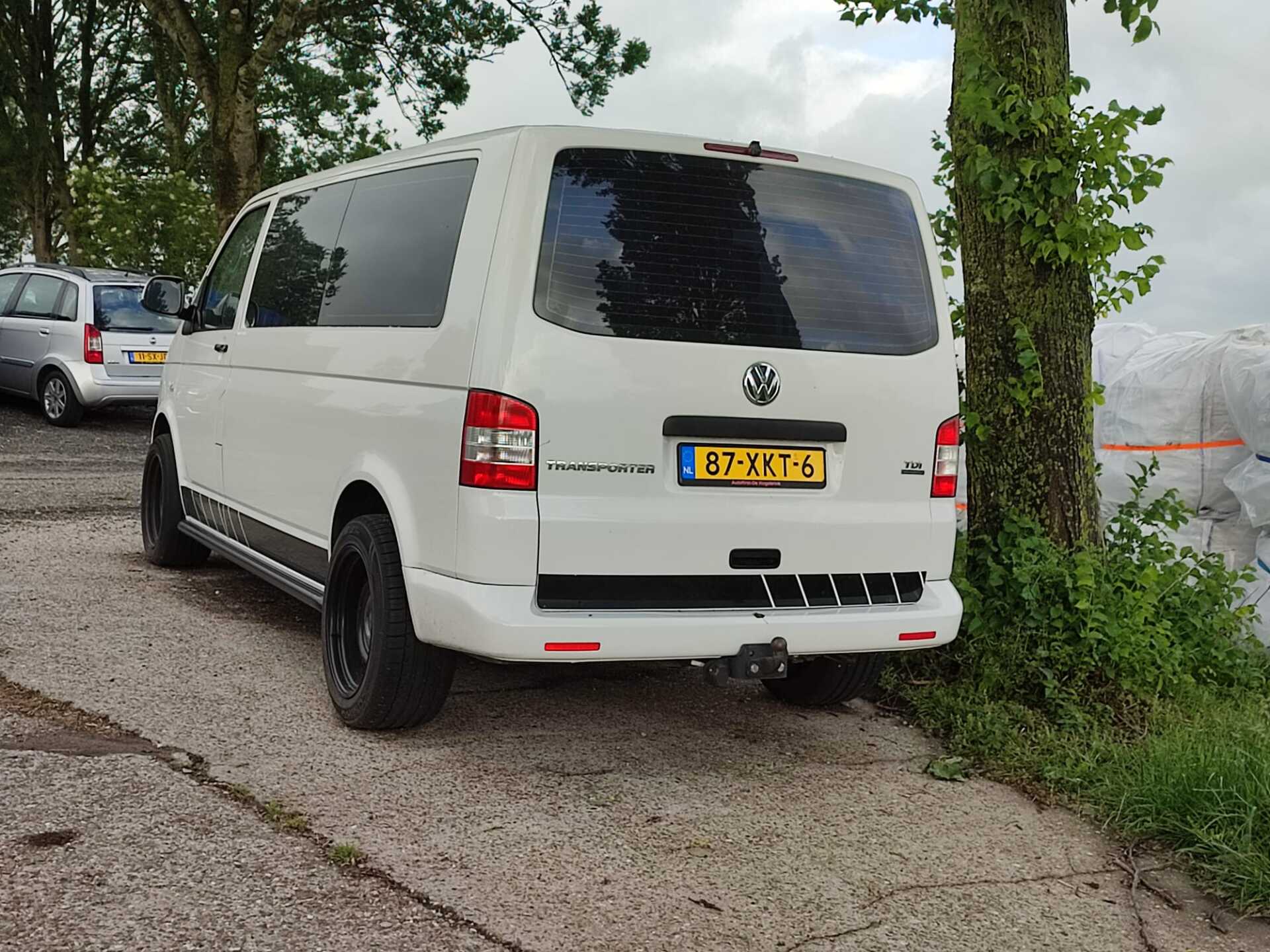 Witte Volkswagen Transporter bestelbus met getinte ramen, geparkeerd op asfalt nabij een boom en andere voertuigen; Nederlands kenteken.