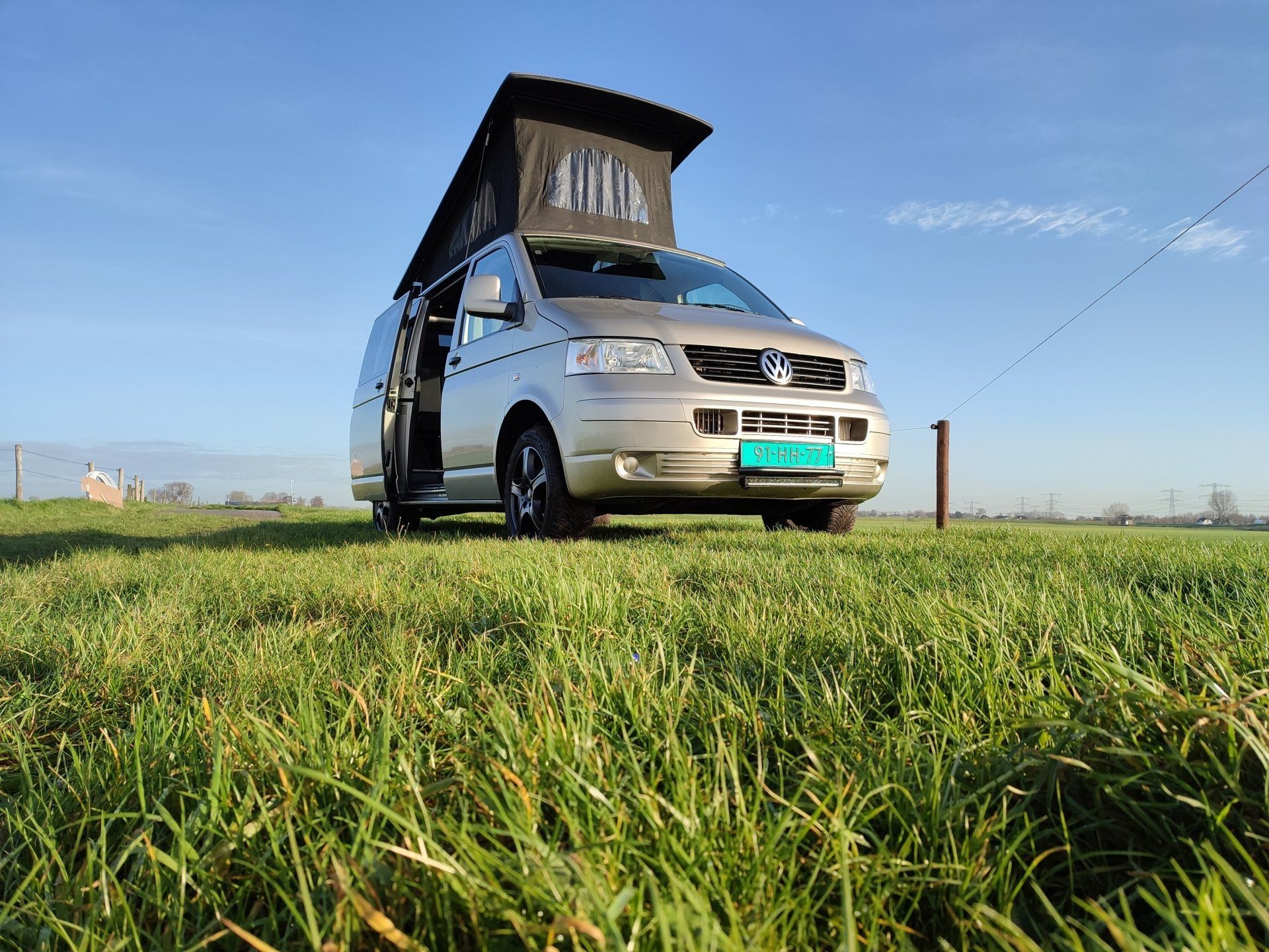 Een zilverkleurige camper met hefdak staat geparkeerd in een grasveld op een zonnige dag.