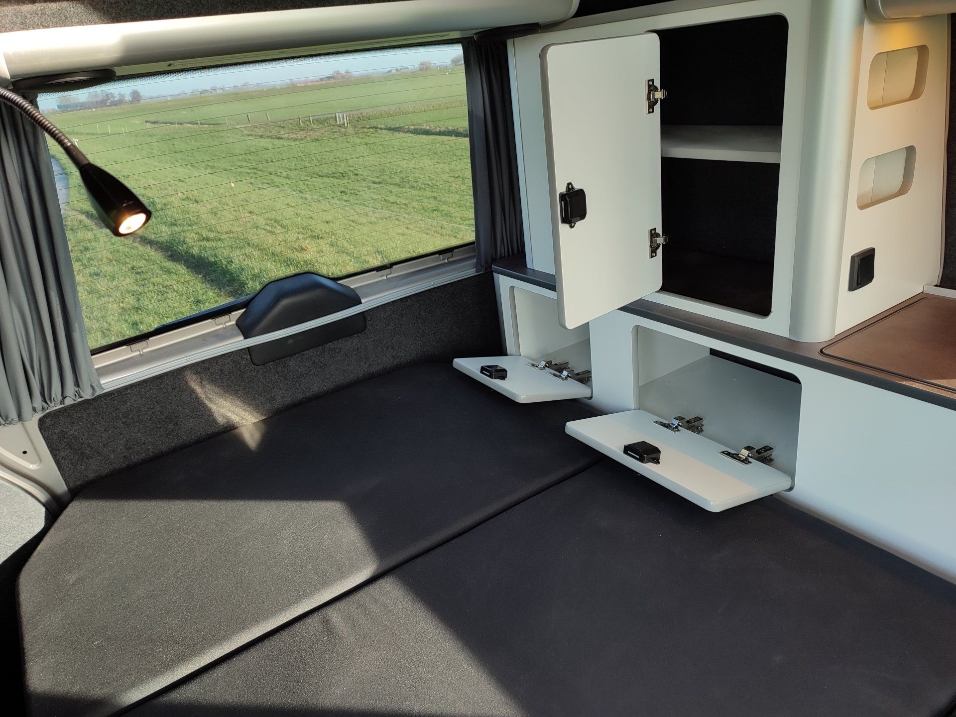 Interieur van een camper met een bed, opbergruimte en uitzicht op een groen veld.