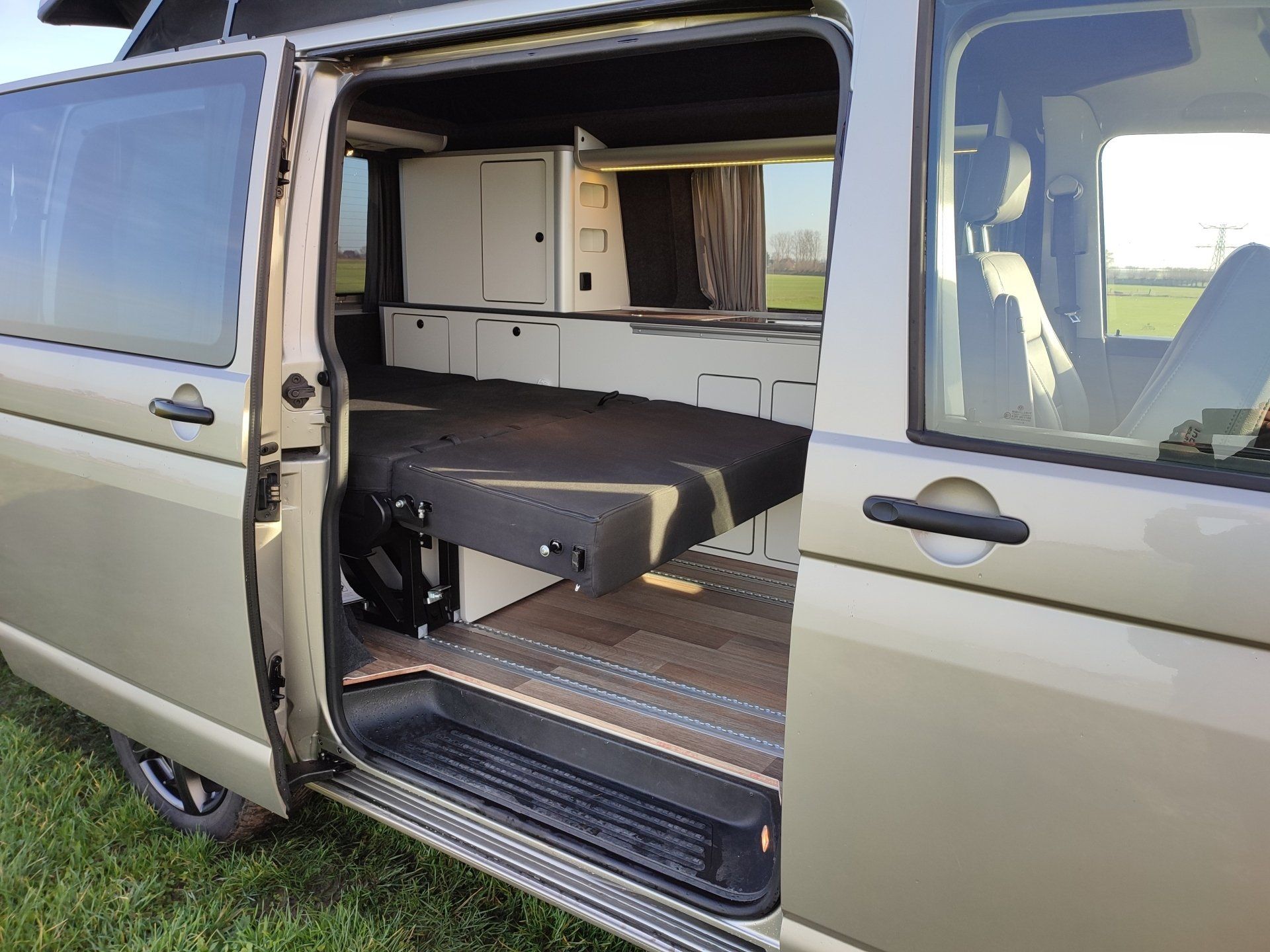 Interieur van een camper, bed uitgeklapt, kastjes zichtbaar. Open deur onthult interieur. Beige buitenkant, dagomgeving.