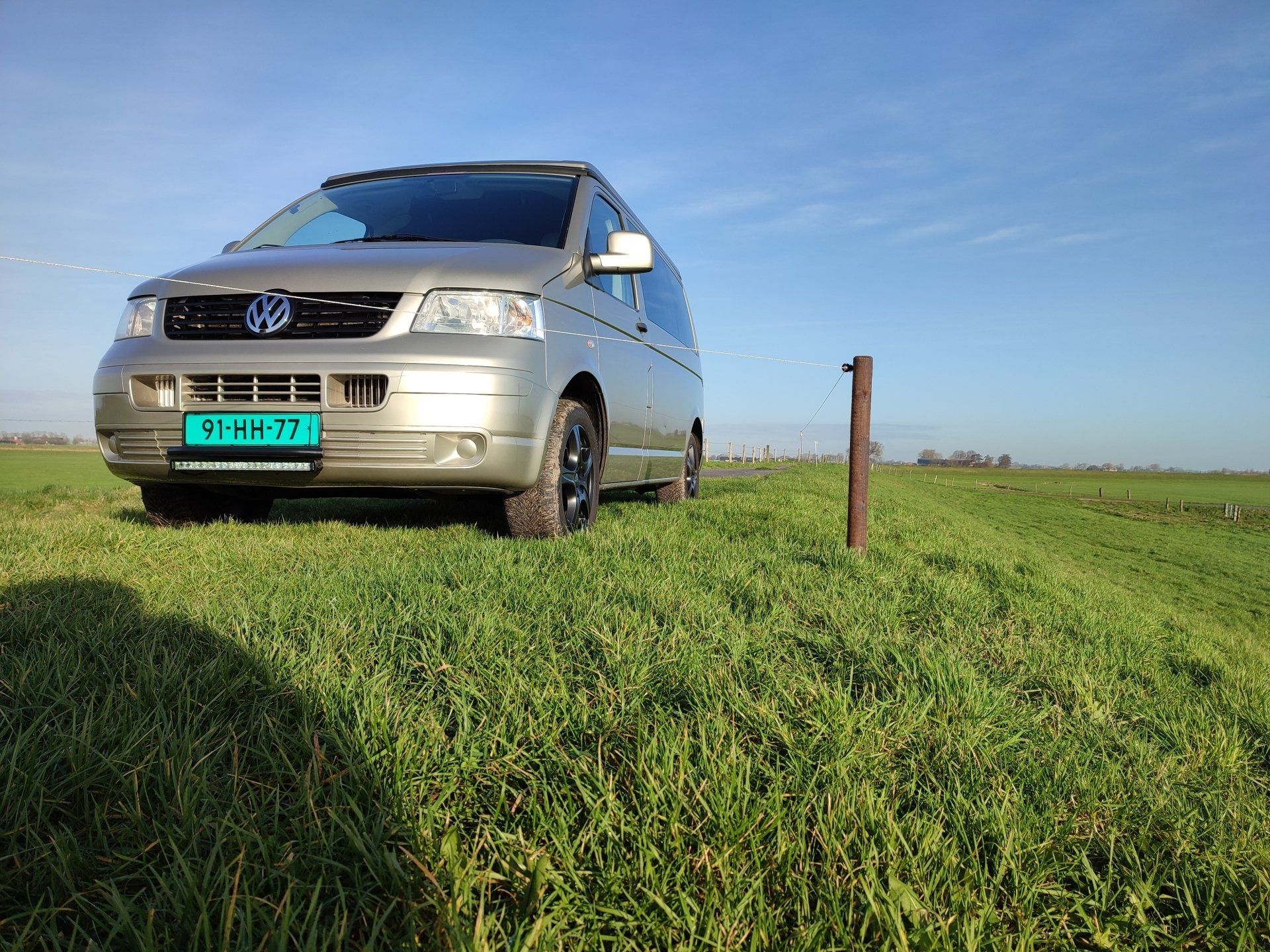 Een zilverkleurige Volkswagenbus staat geparkeerd op een grashelling onder een blauwe hemel, in een landelijke omgeving.