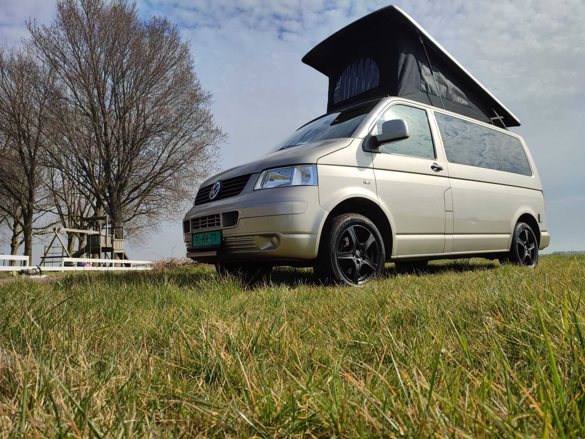 Een beige camper met hefdak staat op een grasveld onder een bewolkte hemel.