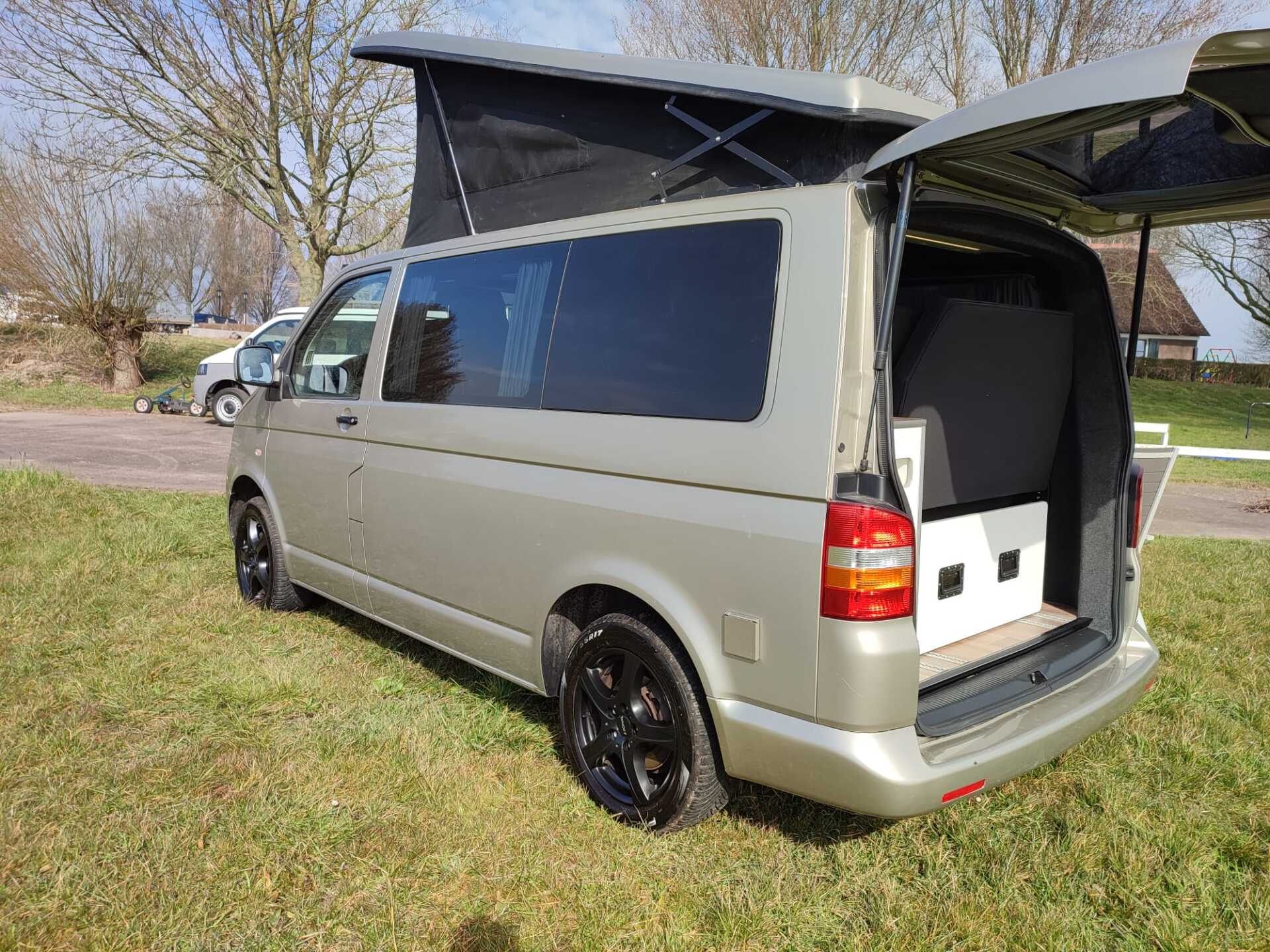 Zilverkleurige camper met hefdak en open achterklep, geparkeerd op het gras.