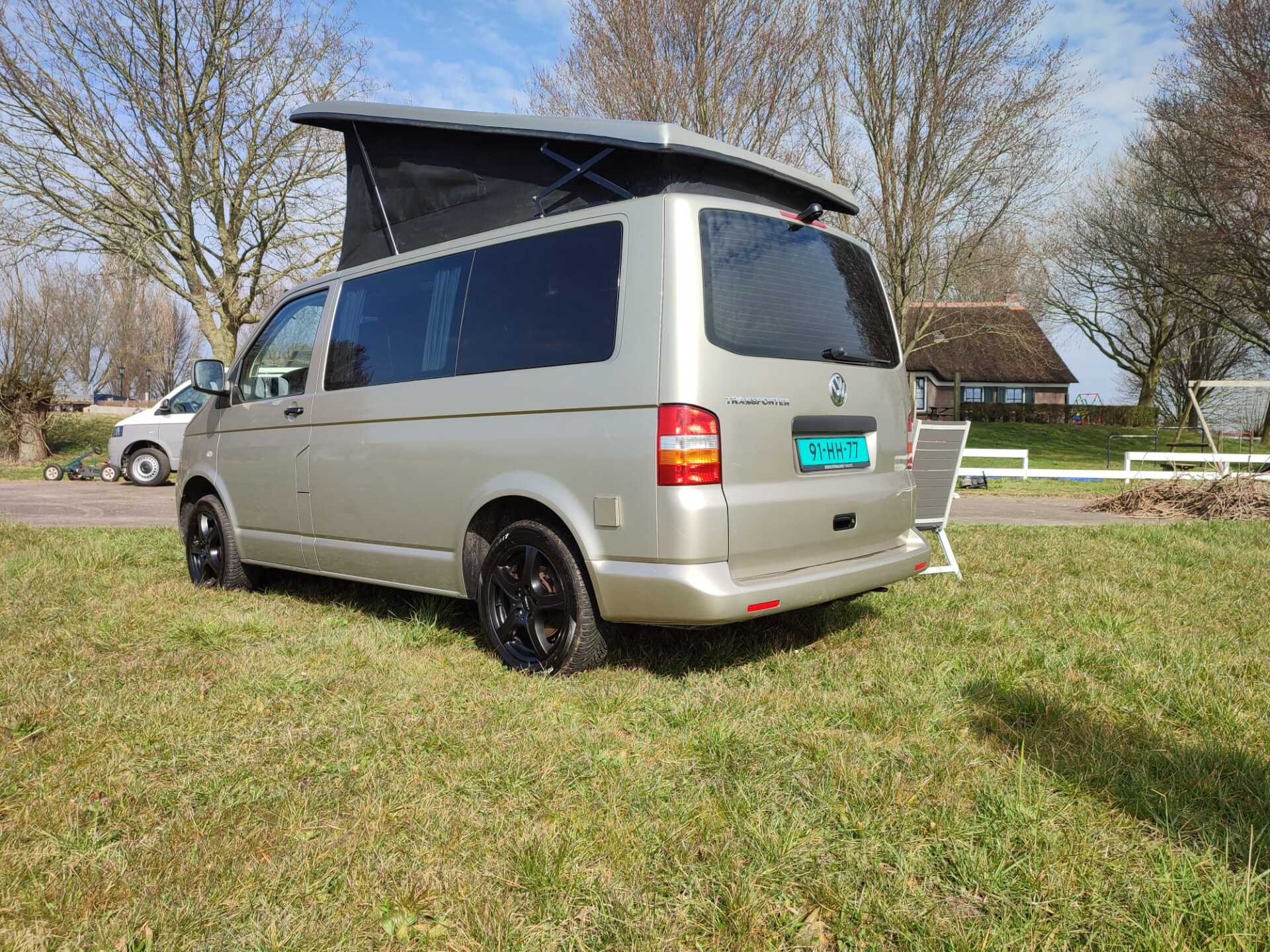 Een goudkleurige camper met een hefdak staat geparkeerd op het gras, vlakbij bomen en een gebouw.