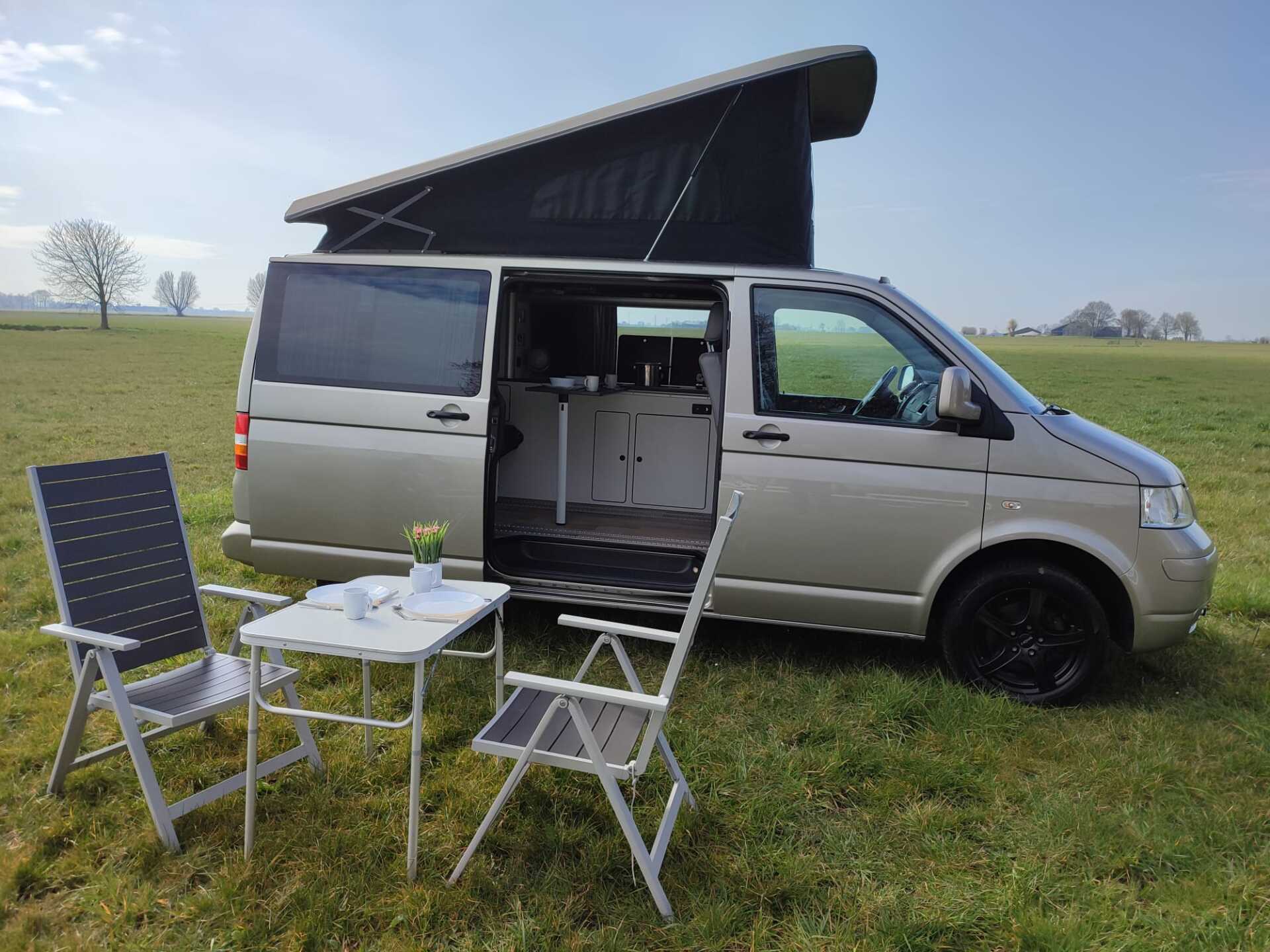 Een beige camperbusje met hefdak staat geparkeerd in een grasveld, met een klein tafeltje en klapstoelen.