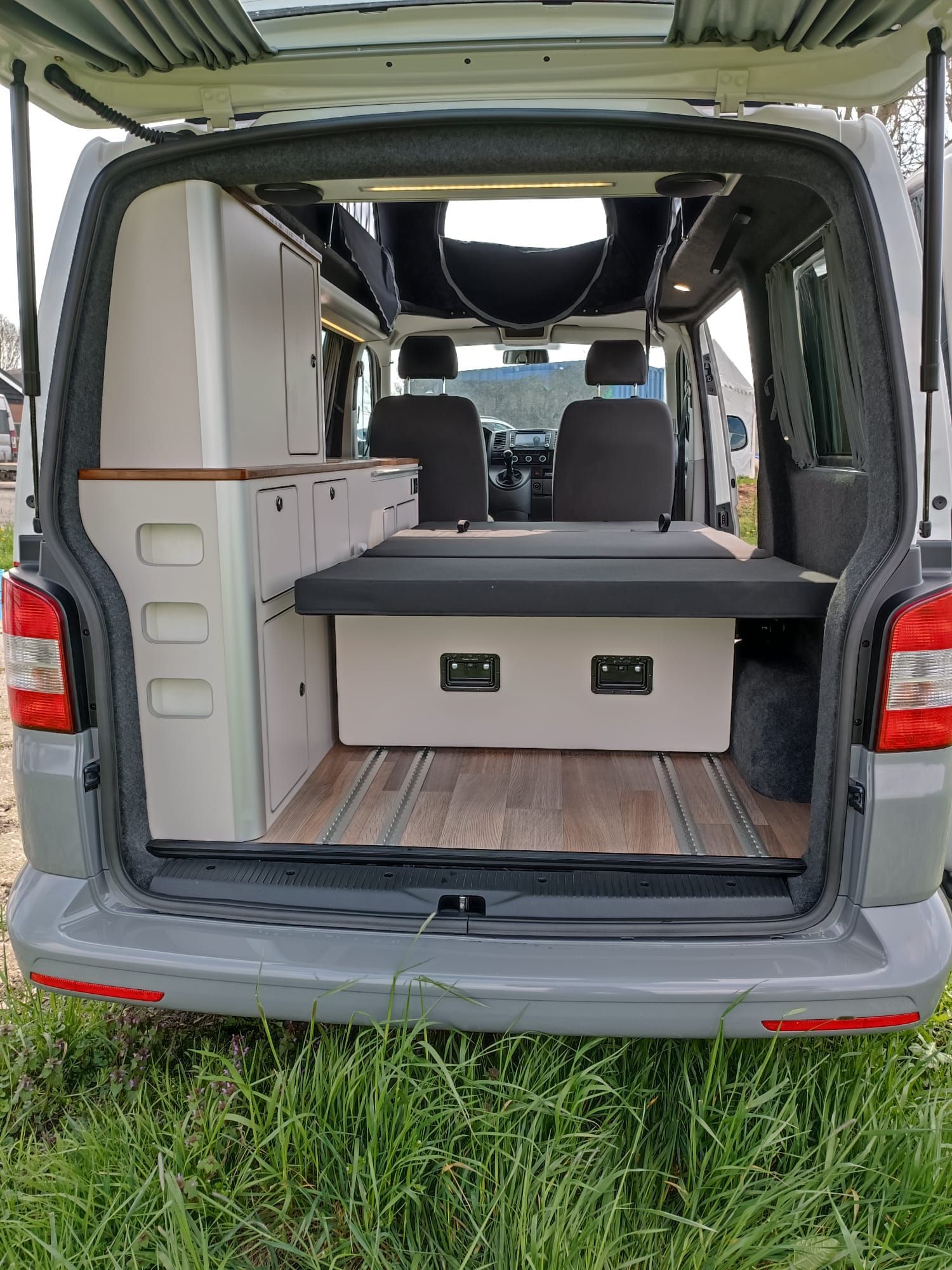 Achteraanzicht van het interieur van een camper: kasten, bed en open dak met natuurhouten vloer.