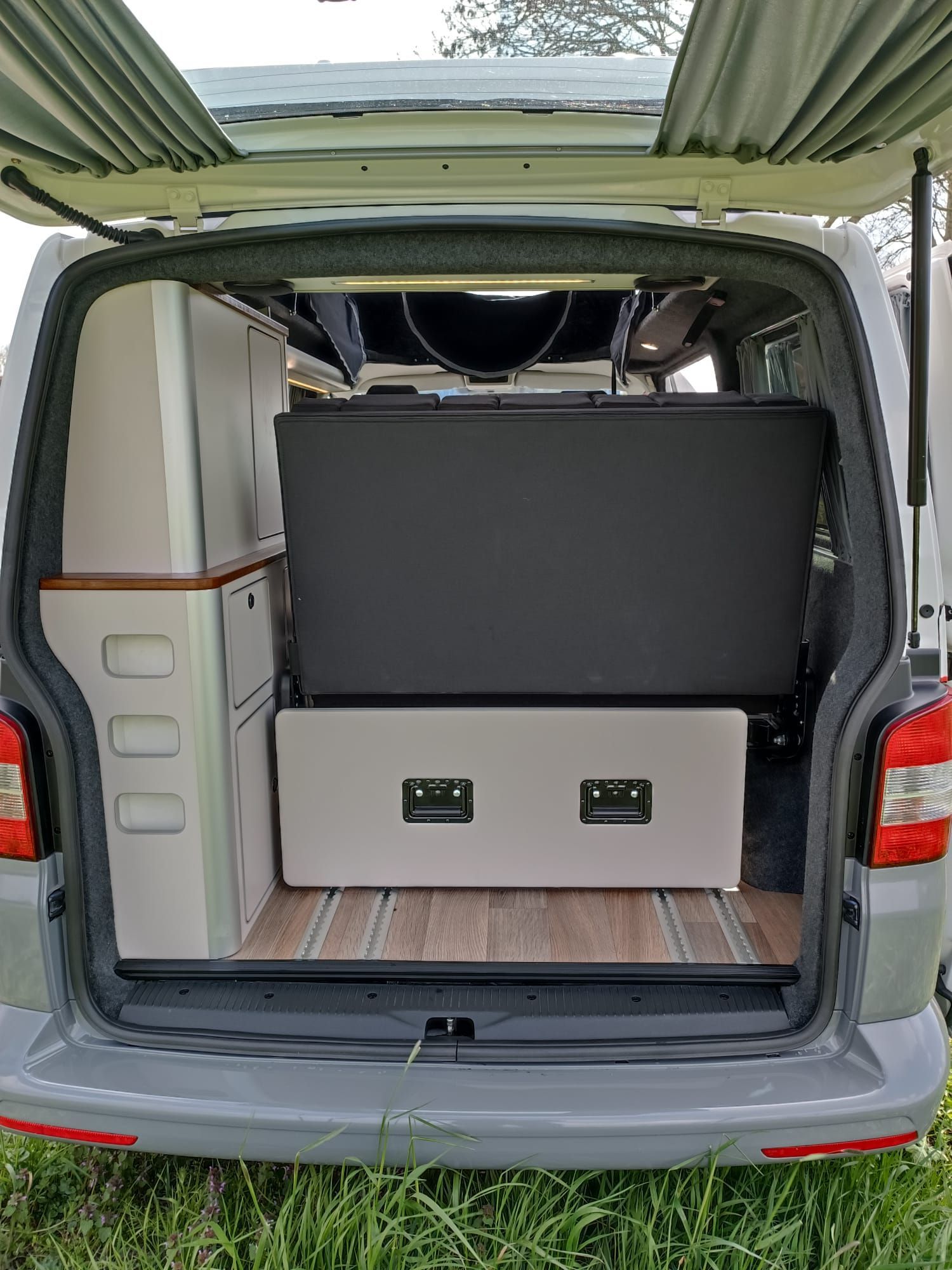 Achterkant van een grijze camper met een bed, kastjes en een open luik.