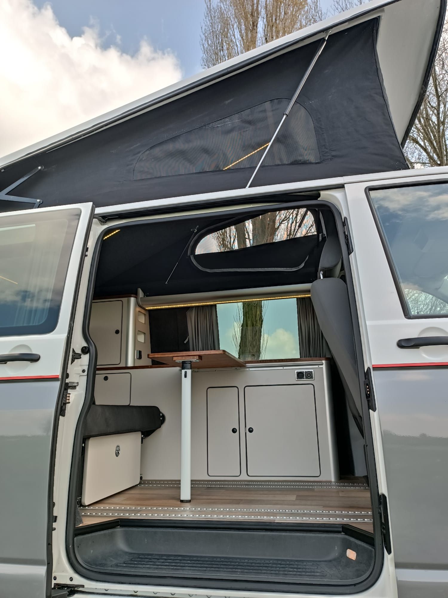 Open deur van een camperbusje, waardoor het interieur zichtbaar is met kastjes, een tafel en een hefdak. Witte en grijze buitenkant.