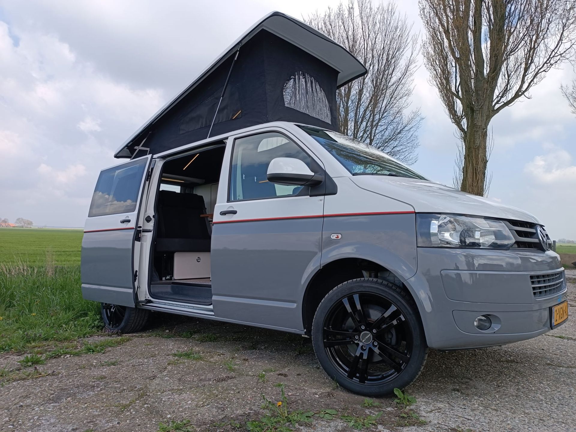 Grijze en witte camper met hefdak en open zijdeur geparkeerd op grind, met een veld en een boom op de achtergrond.