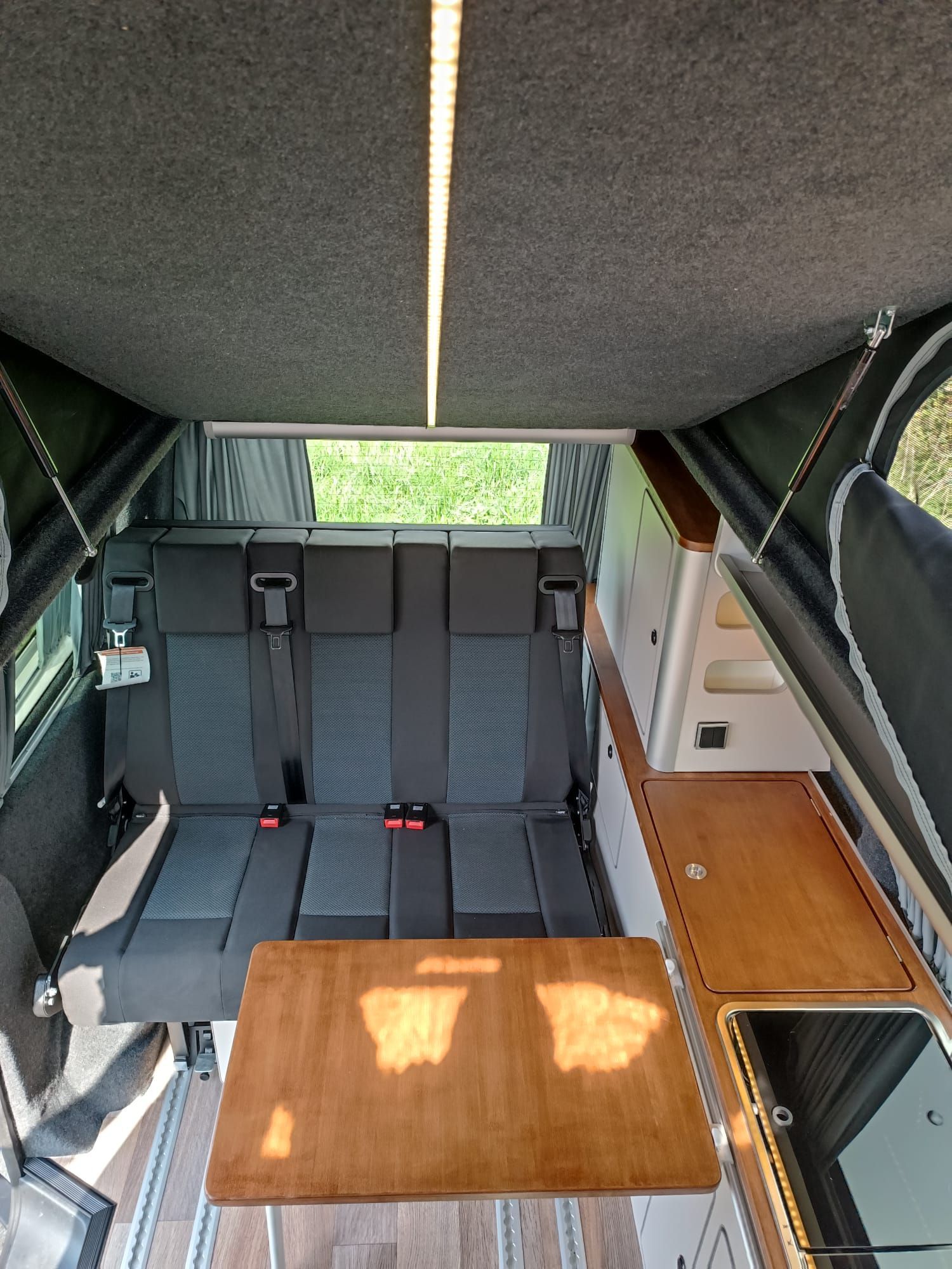Interieur van een camper met een bank, tafel en keukenhoek. Grijze en houttinten overheersen.