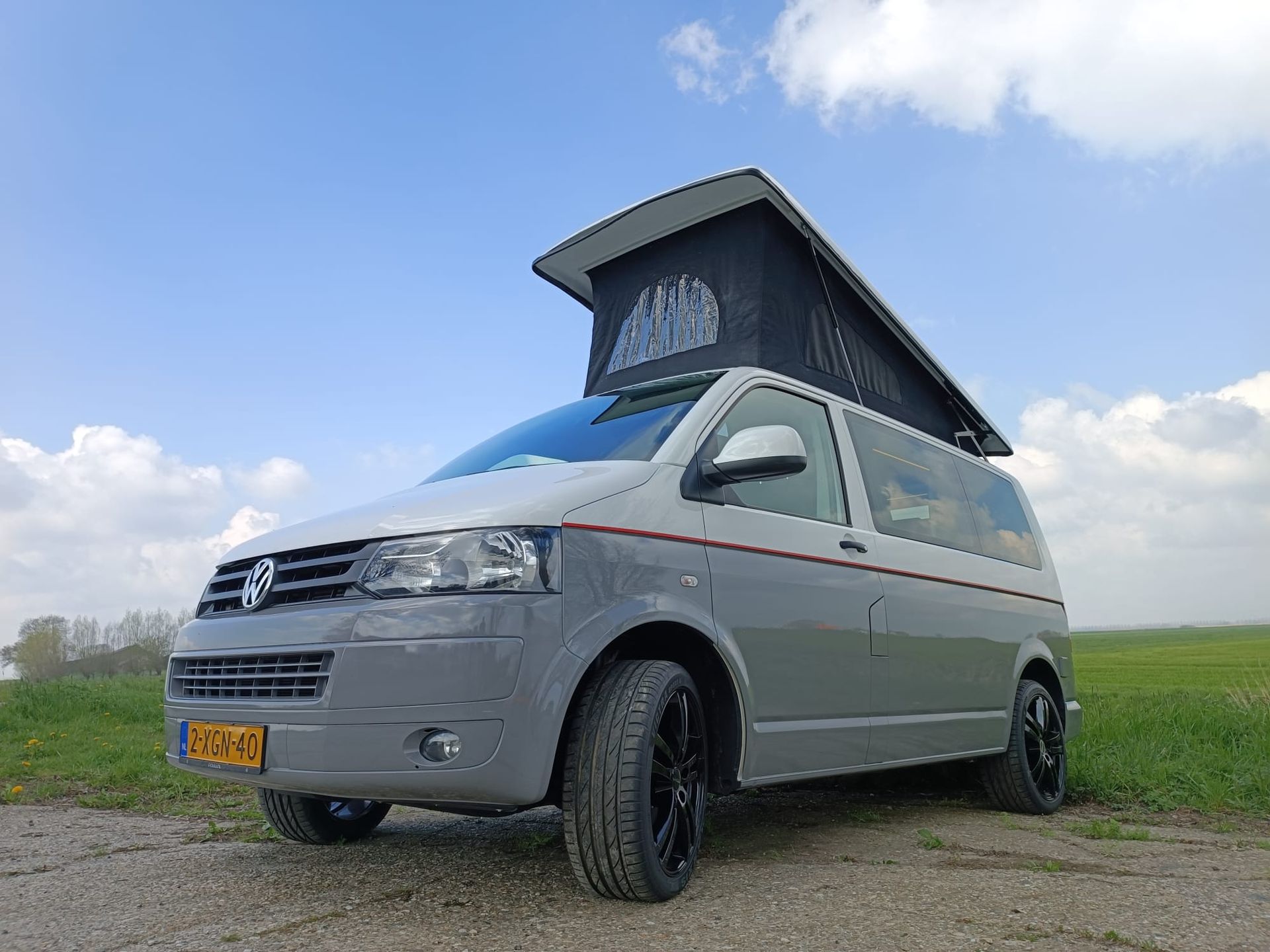 Een grijs-witte camper met hefdak staat geparkeerd op een onverharde weg onder een blauwe hemel.