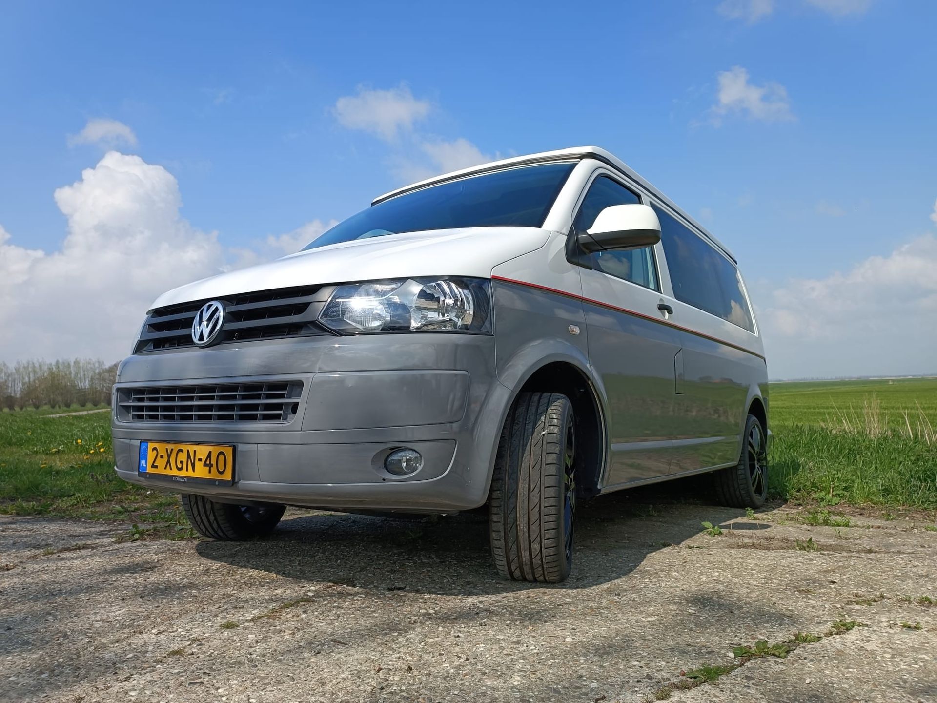 Grijze en witte Volkswagen camperbus geparkeerd op een onverharde weg, zonnige lucht.