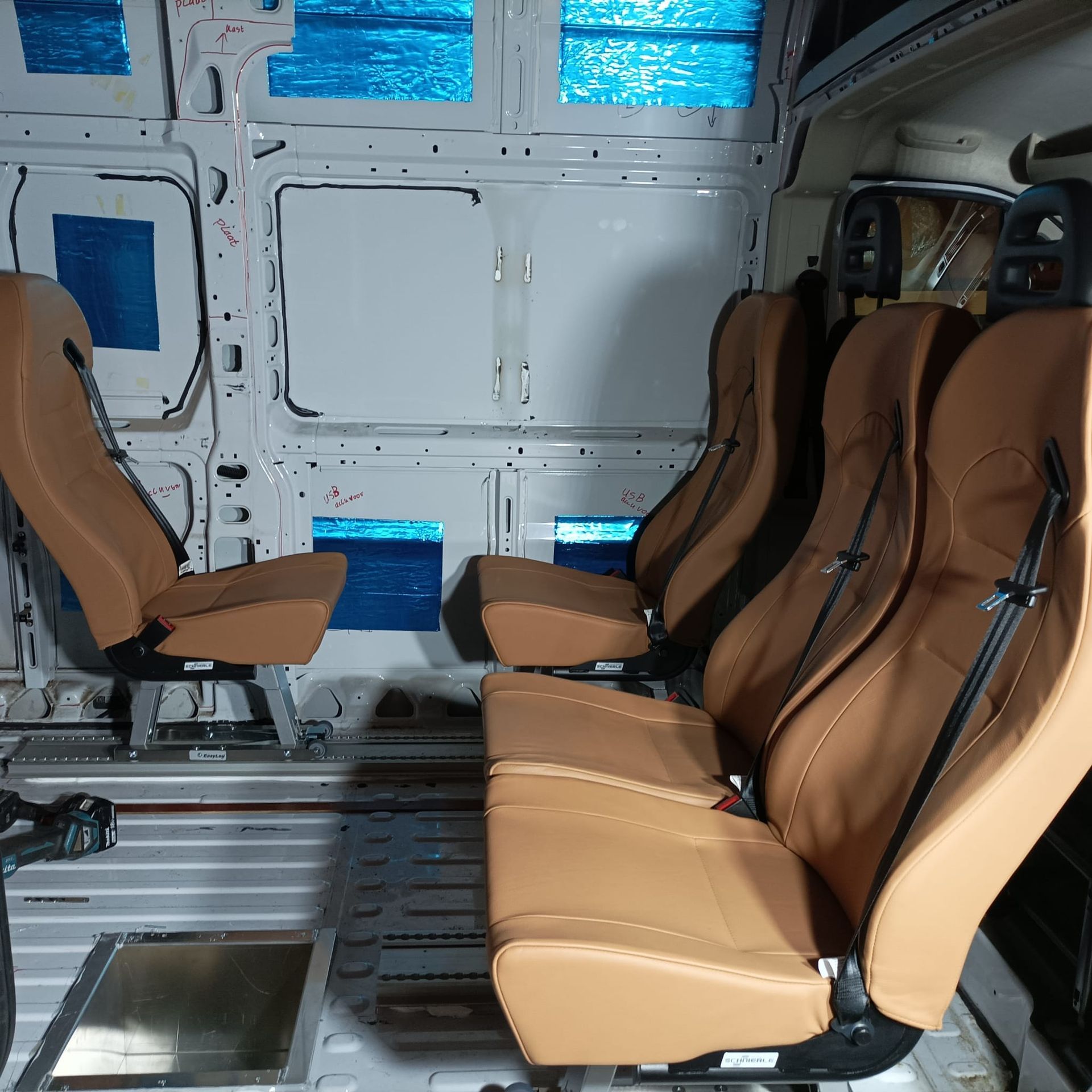 Beige lederen stoelen in een voertuig met witte wanden, waarschijnlijk een bestelbus. Blauw materiaal zichtbaar in de wanden.