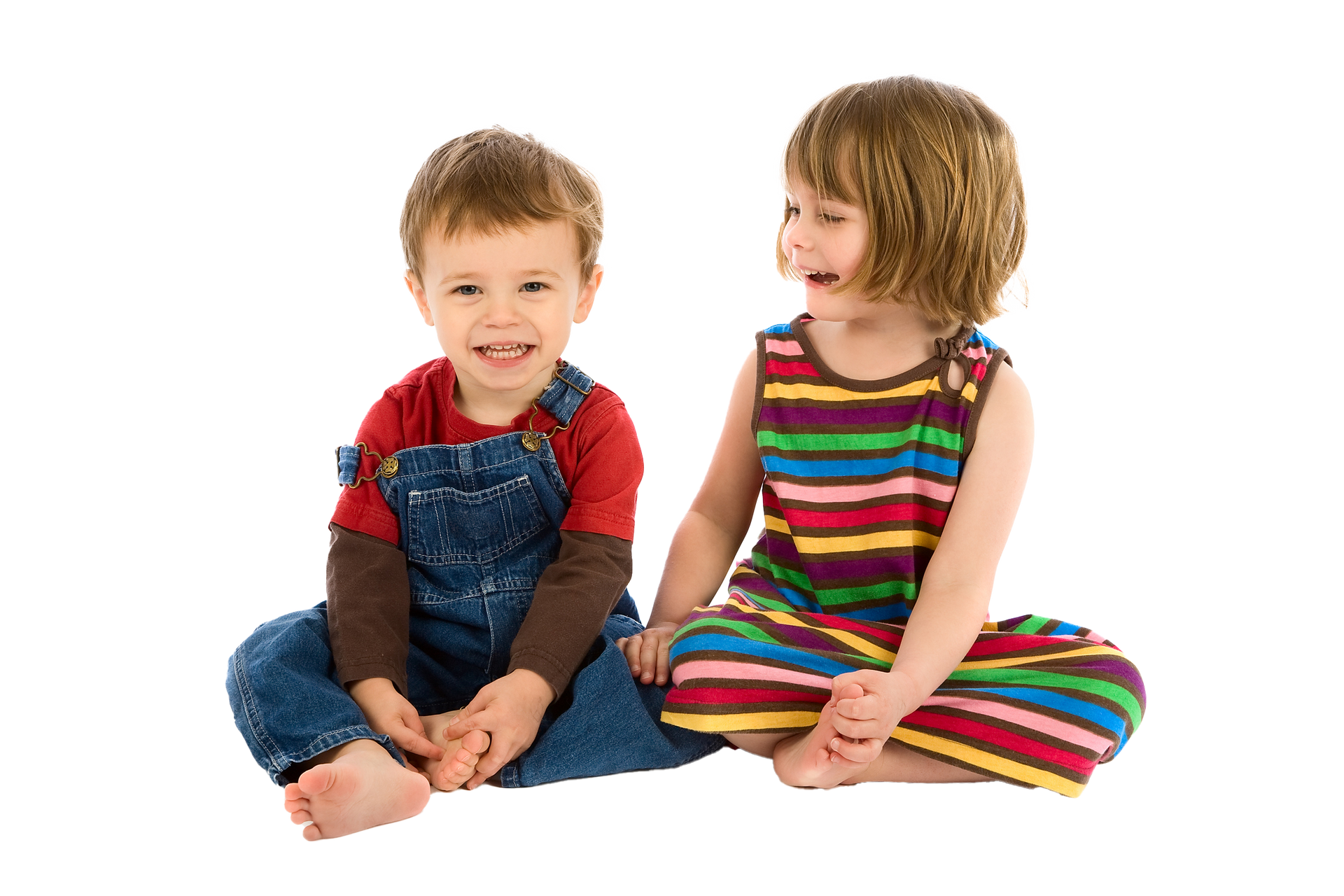 Two young children sitting close together, smiling at the camera. One wears a striped dress; the other has overalls.
