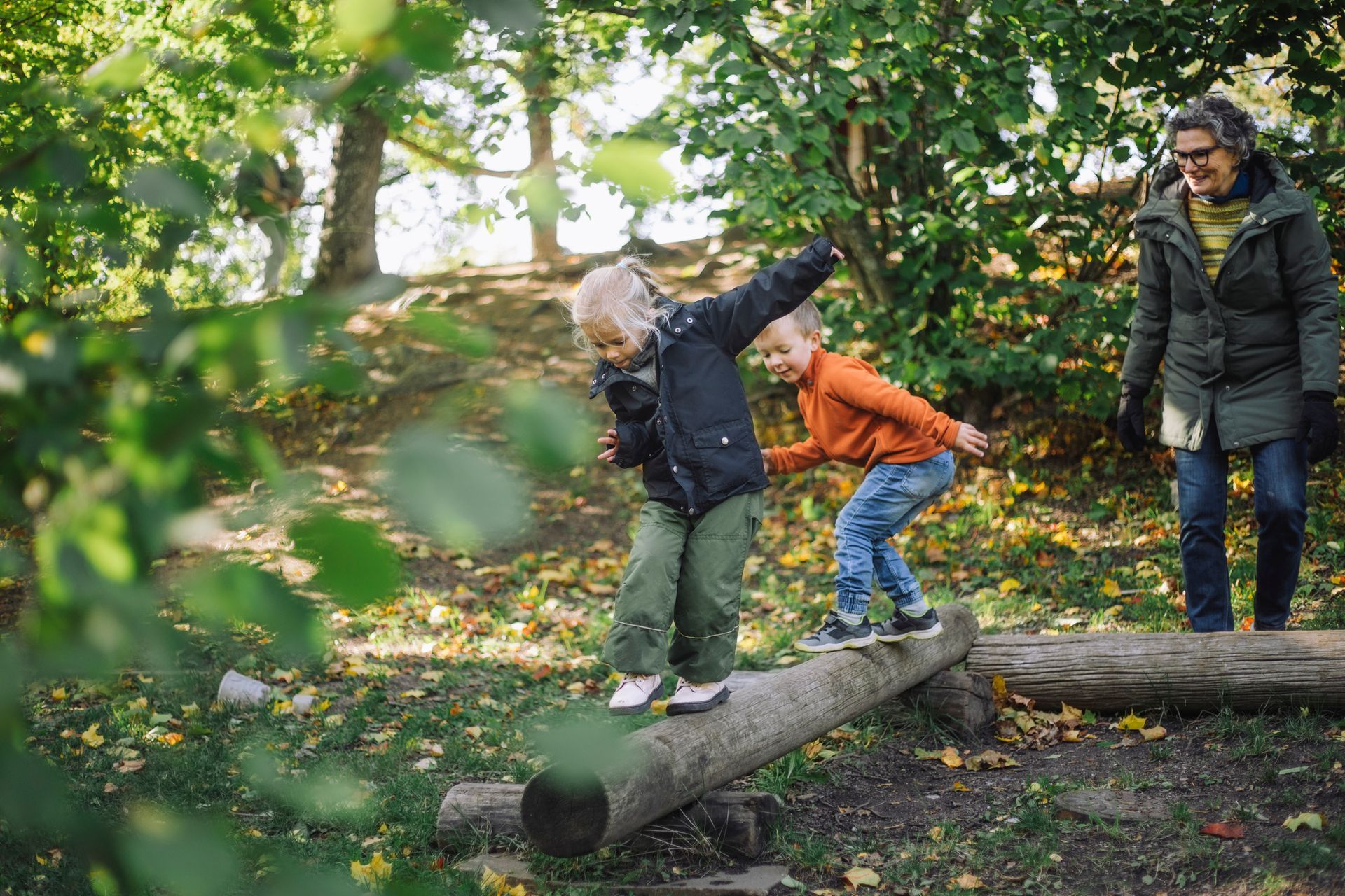 Two children balance on a log in a park as an adult watches.