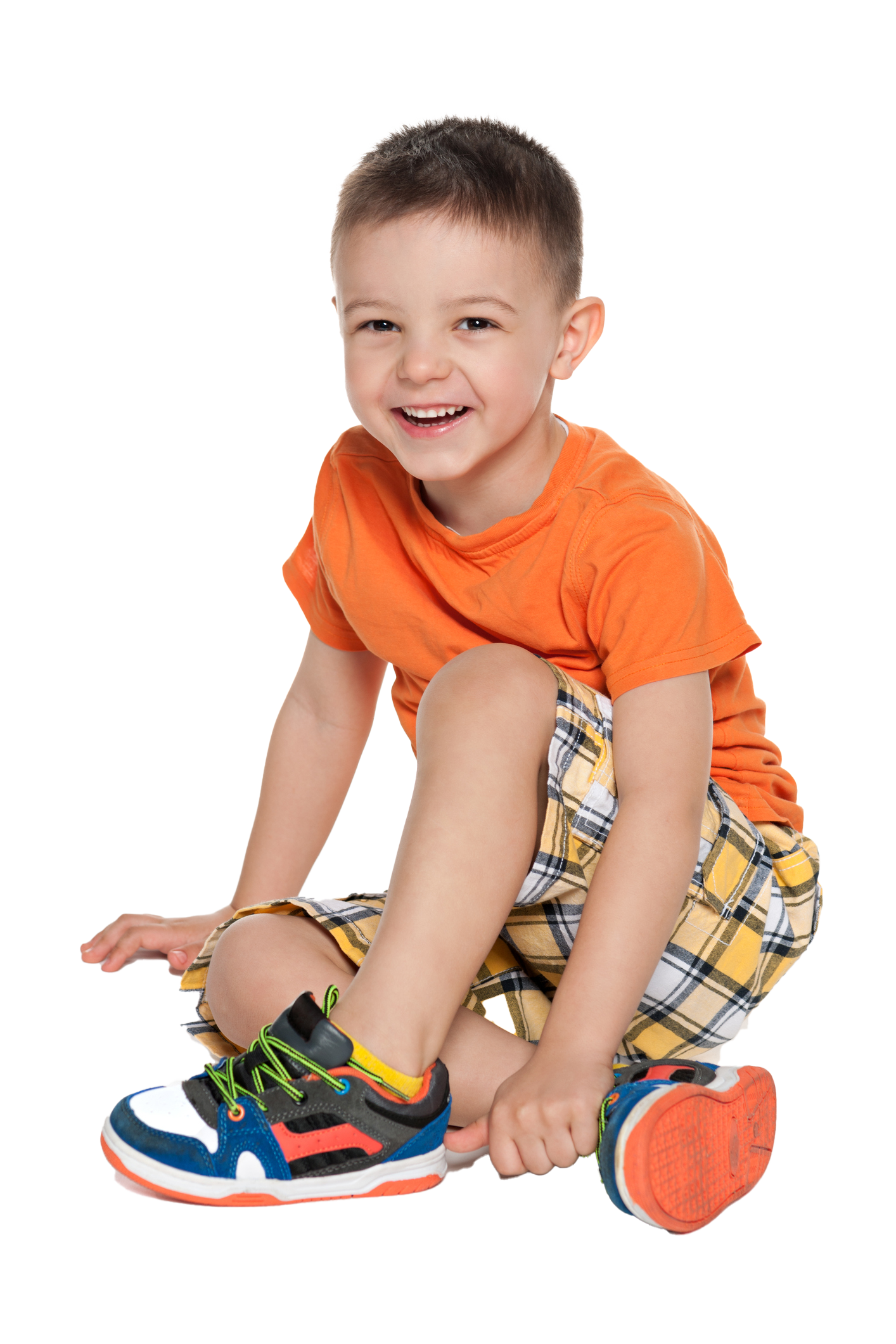 Smiling child in orange shirt and plaid shorts sits on the floor, next to a shoe.