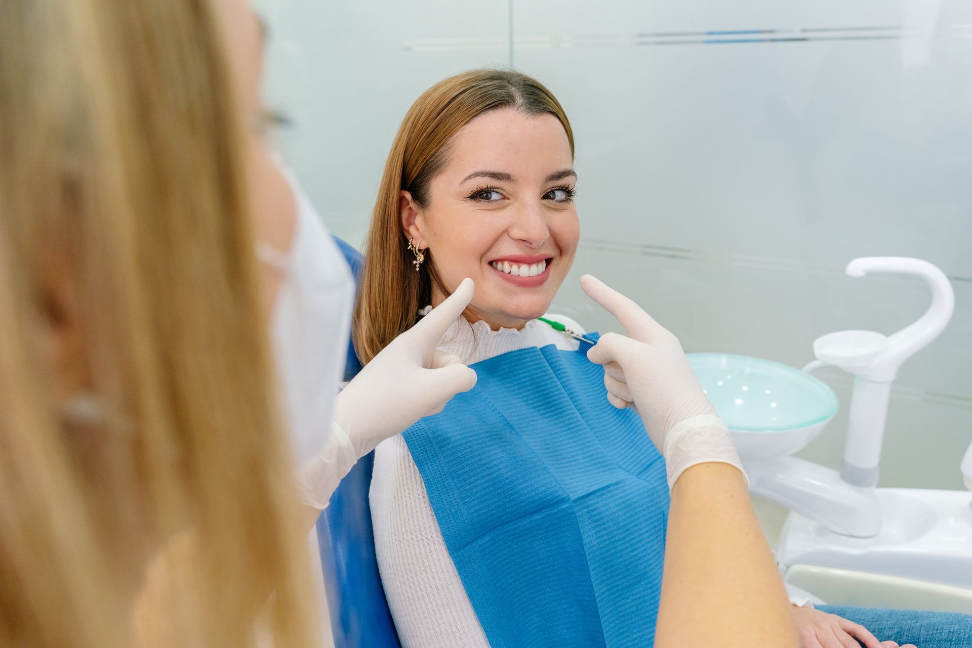 A smiling woman sitting in a dental chair while a dentist in white gloves points toward her teeth.