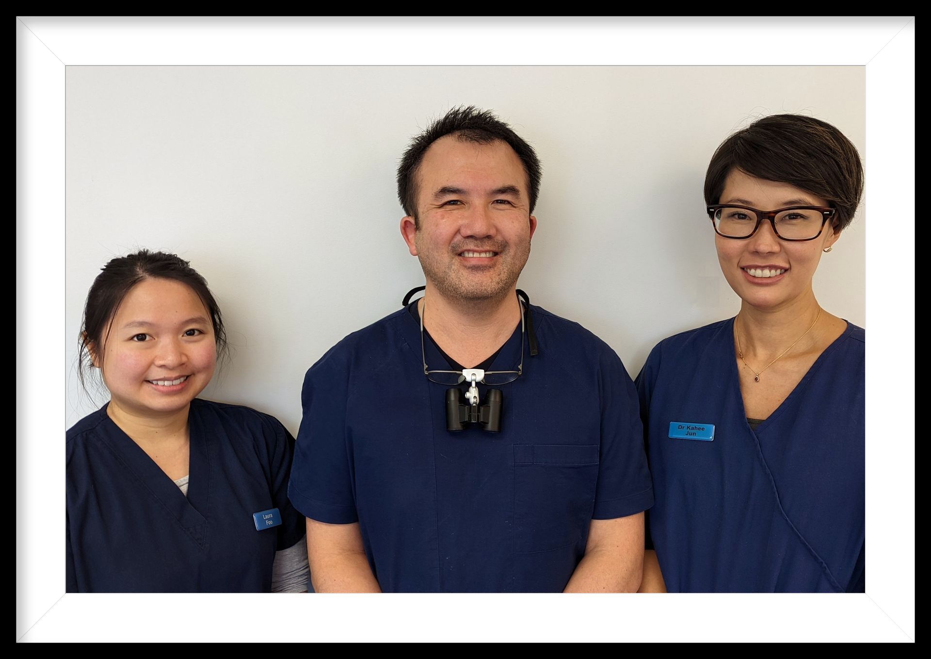 Three dental professionals in blue scrubs smiling in front of a white wall.