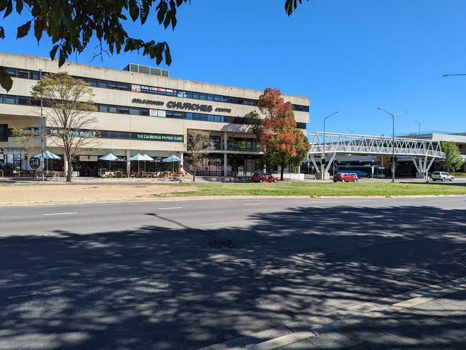 Multi-story building with awnings, pedestrian bridge, trees, and a sunny sky.