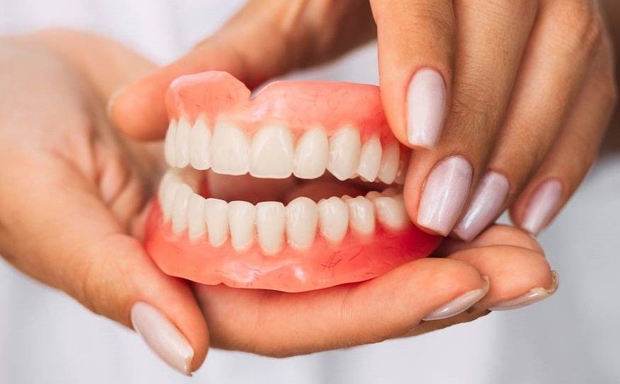 Hands holding a set of complete dentures, pink gums and white teeth.