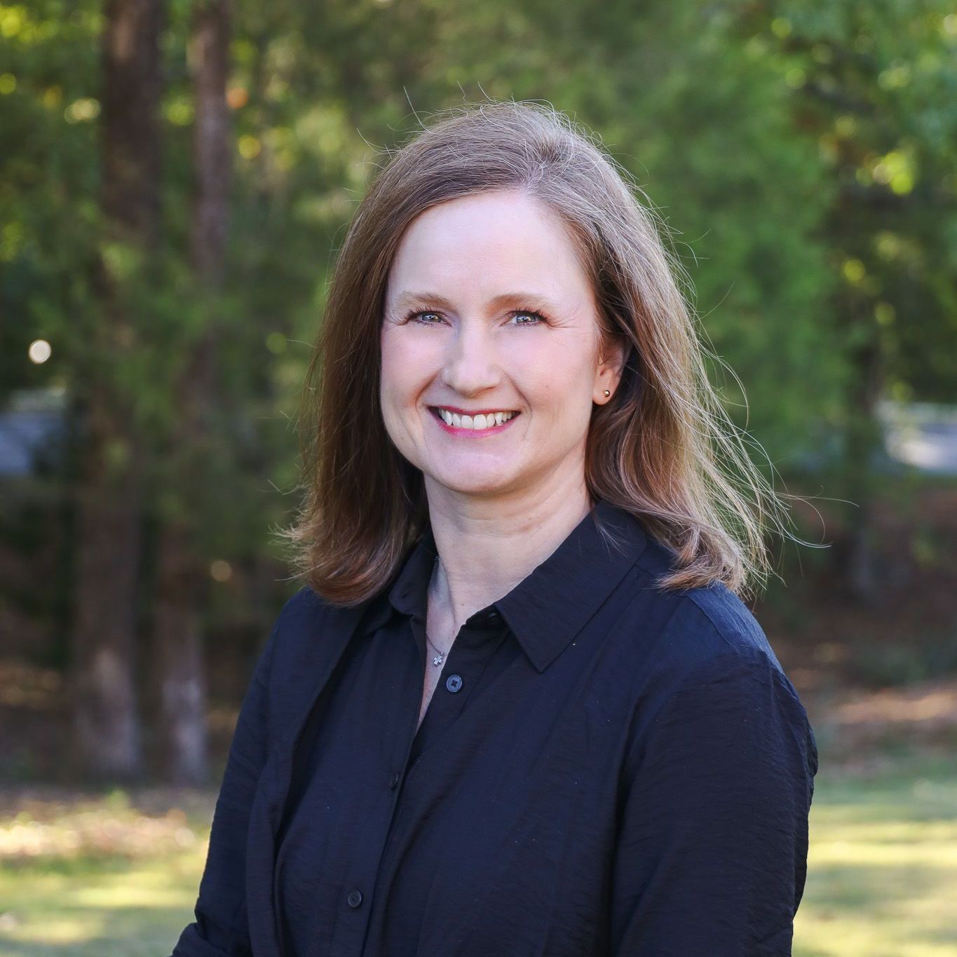 Smiling woman in a dark shirt standing outdoors with trees in the background
