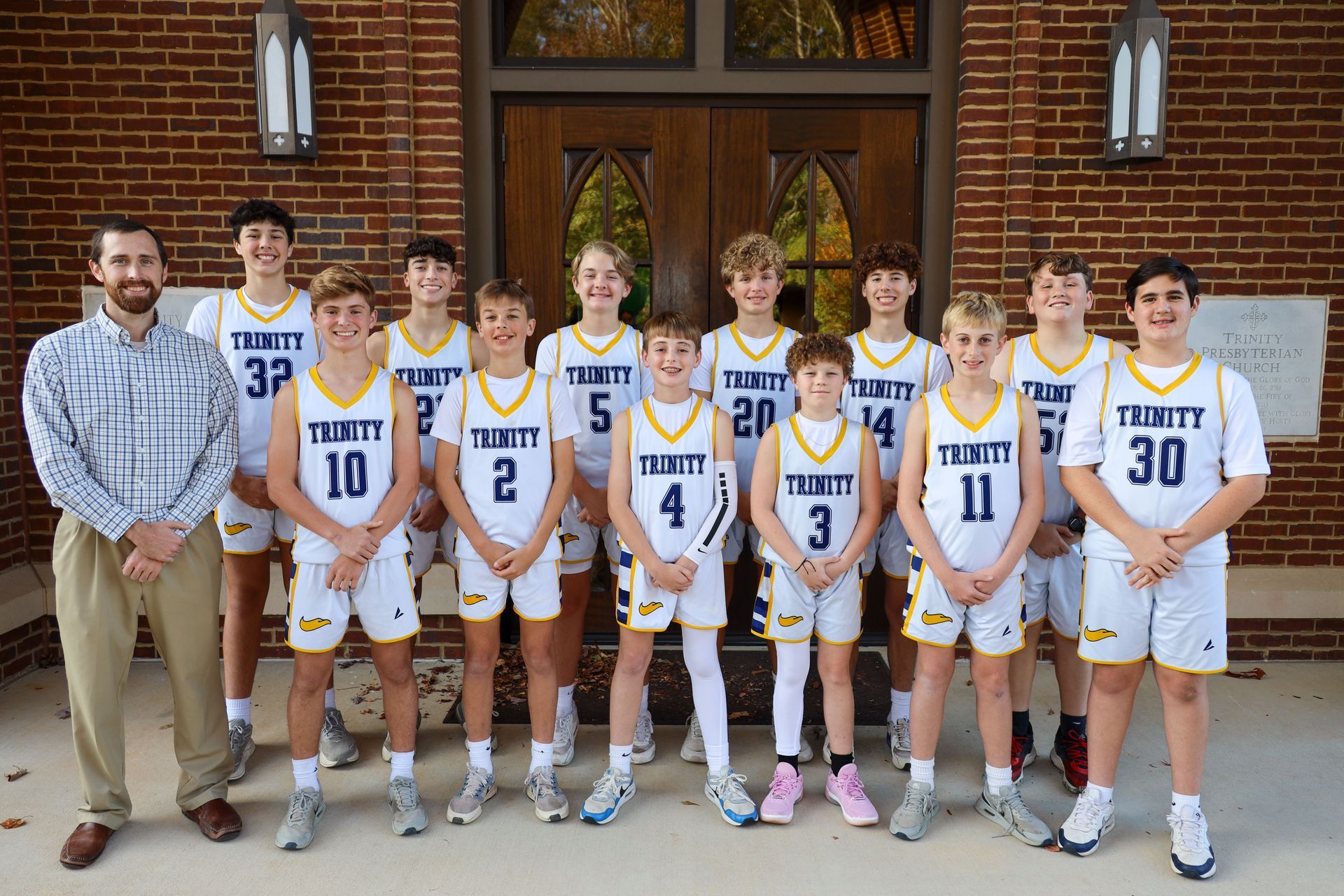Six basketball players and two coaches pose in white Trinity uniforms in front of a building.