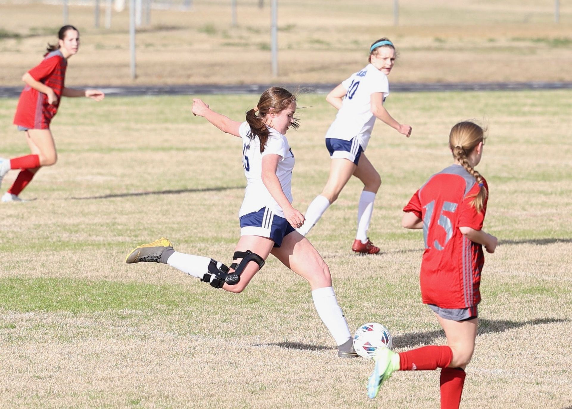 Girls playing soccer on a grassy field, with one in white running toward the ball and others in red nearby