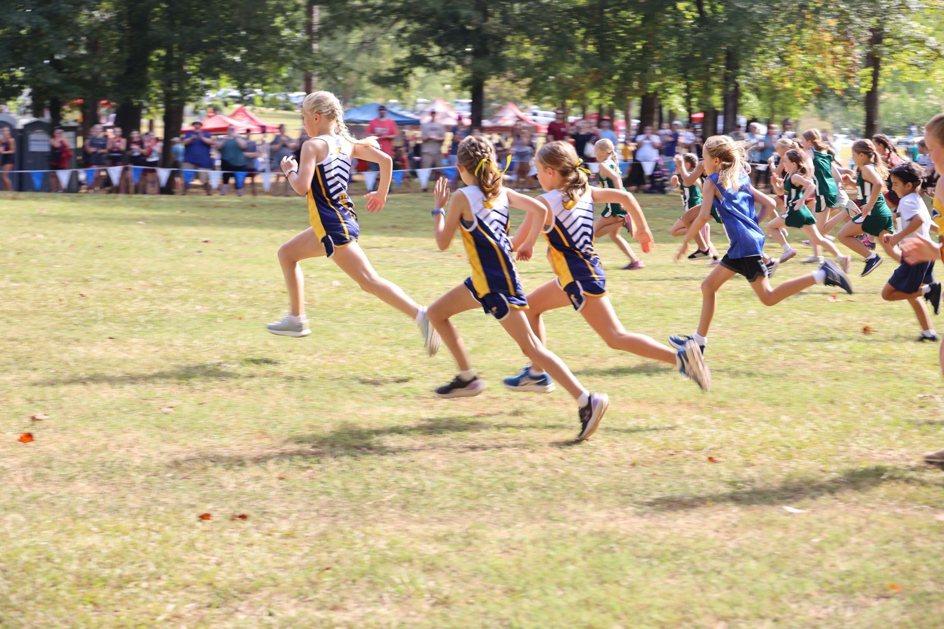 Runners sprint off the start line in a grassy outdoor race with spectators watching in the background