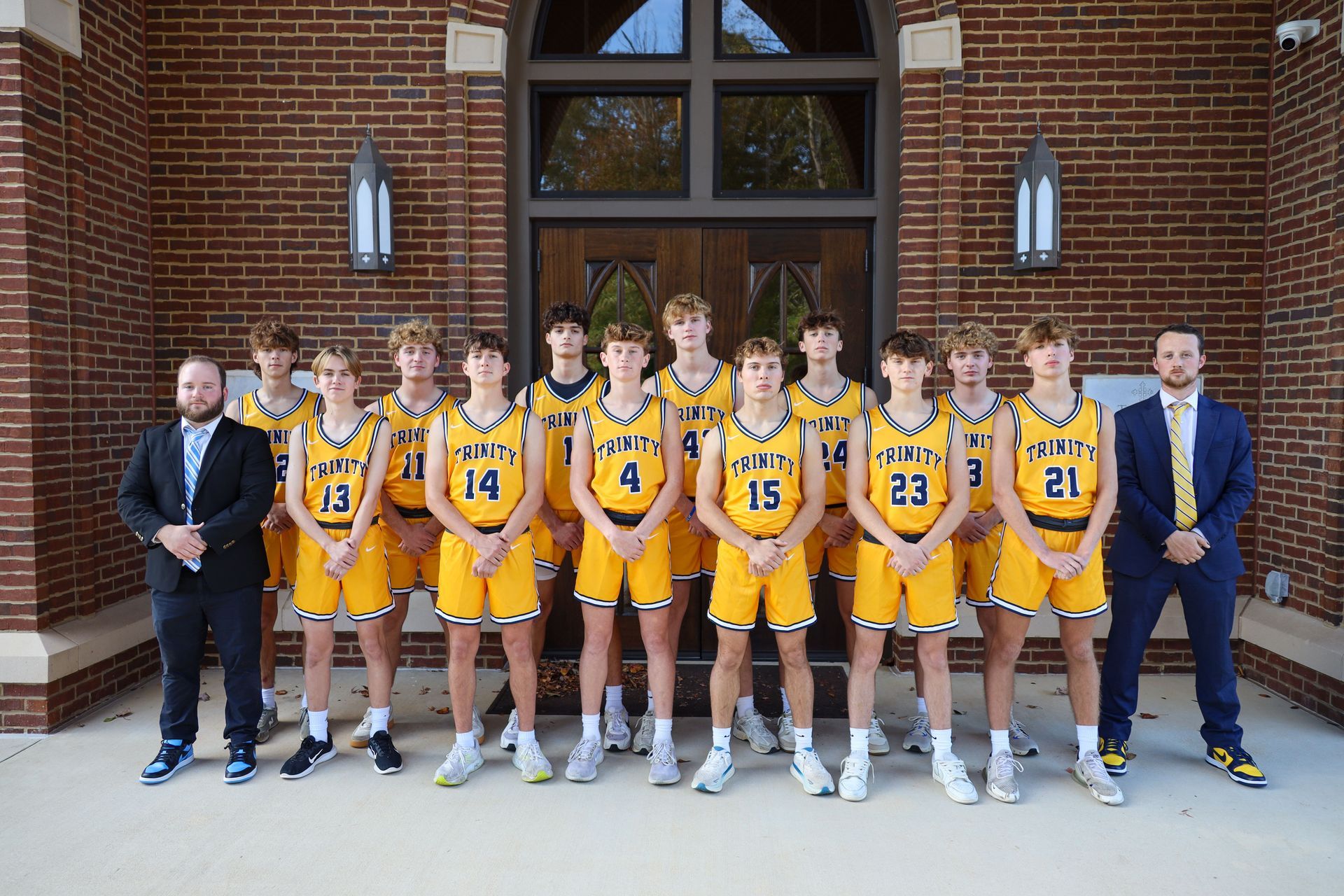 Basketball team in gold uniforms posing with coaches outside a brick building