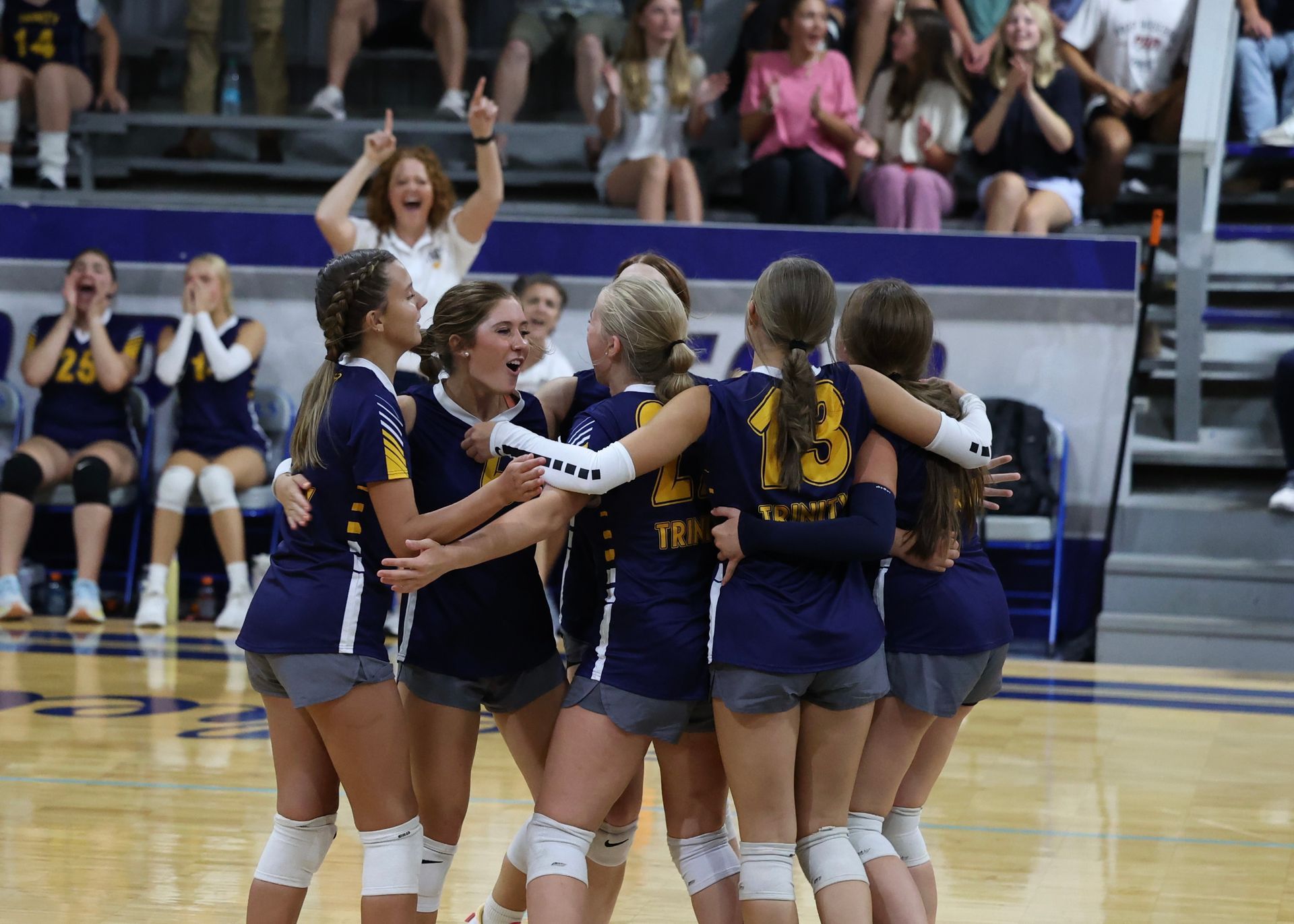 Volleyball team in navy uniforms huddles together on the court after a point, with cheering spectators behind.