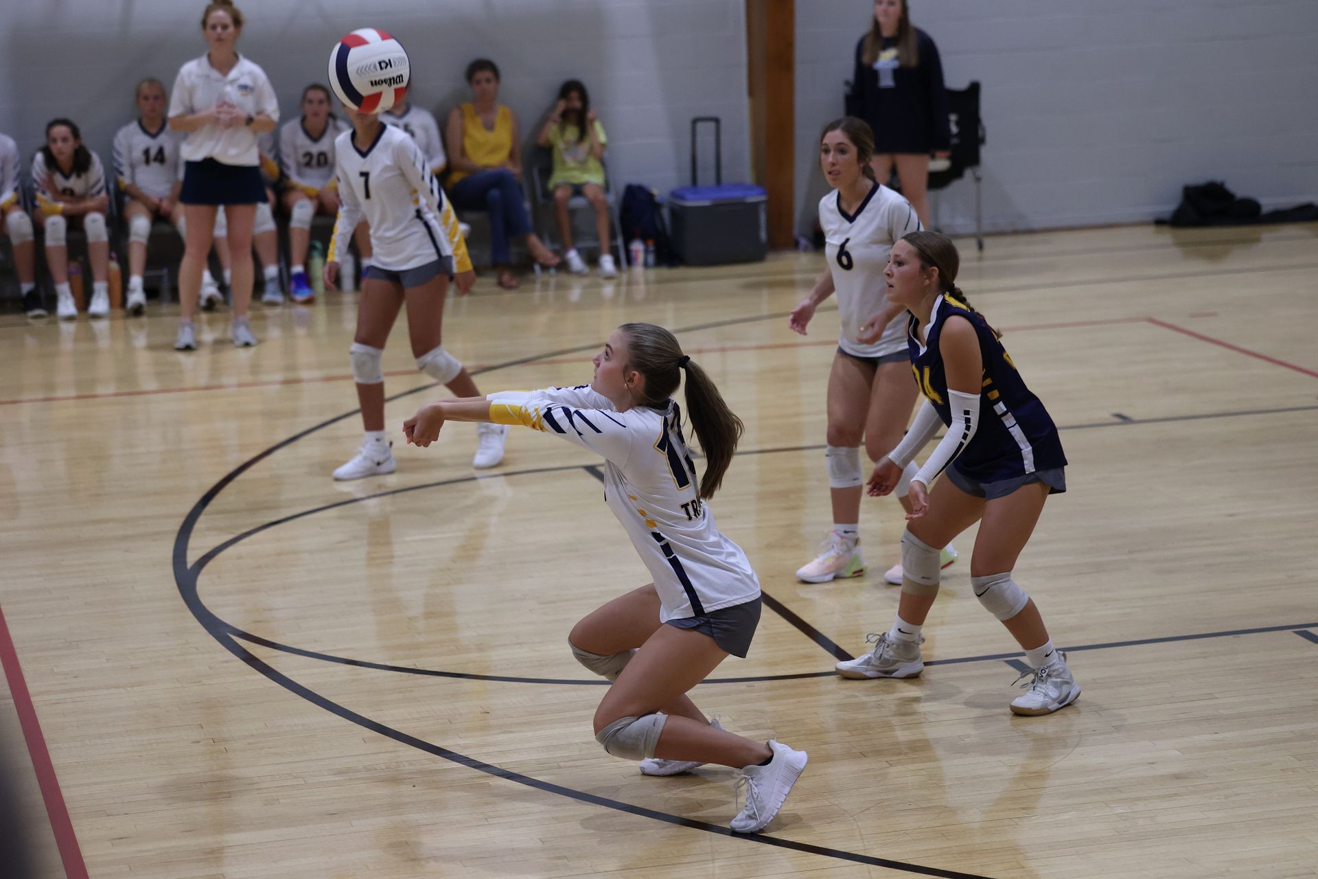 Indoor volleyball game with players in white and dark uniforms, one kneeling to pass the ball