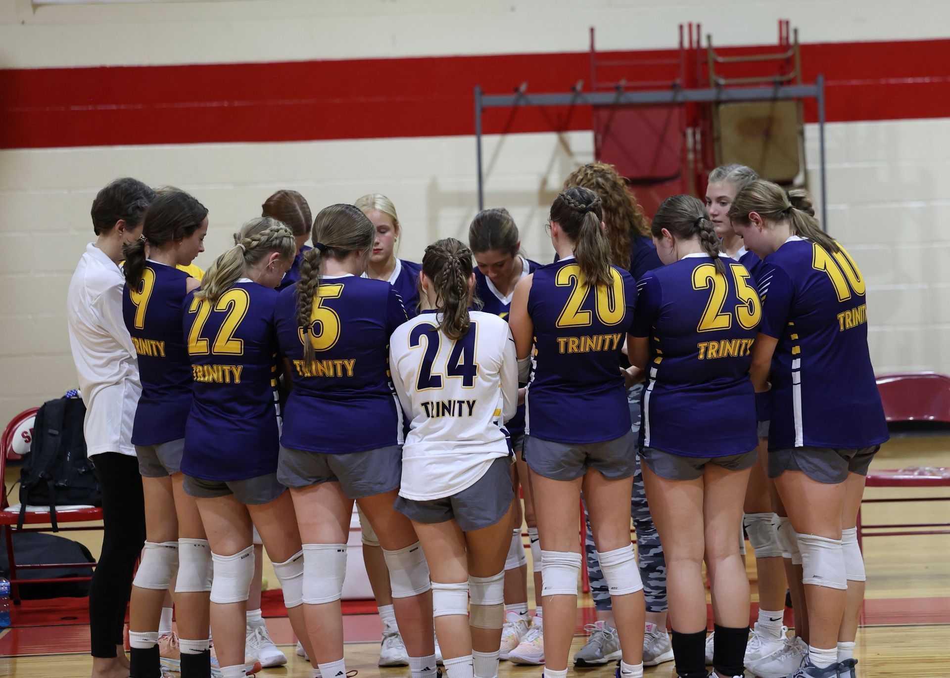 Volleyball team huddling on court in purple and white uniforms, numbers visible on backs
