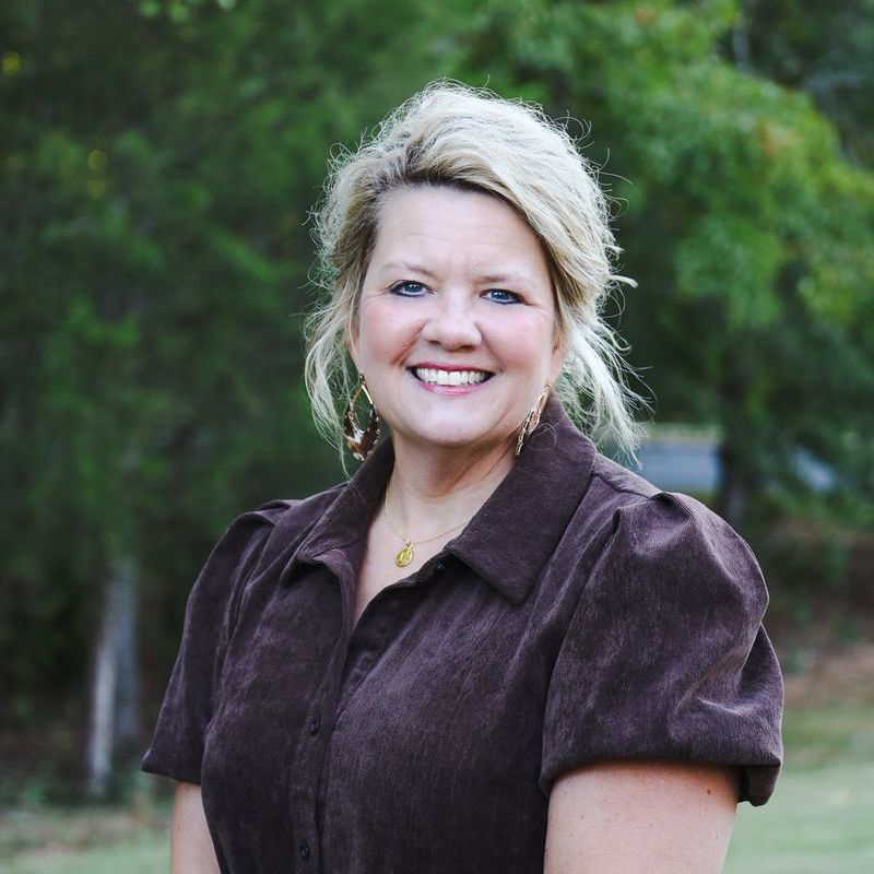Smiling woman in a dark blouse standing outdoors with green trees in the background