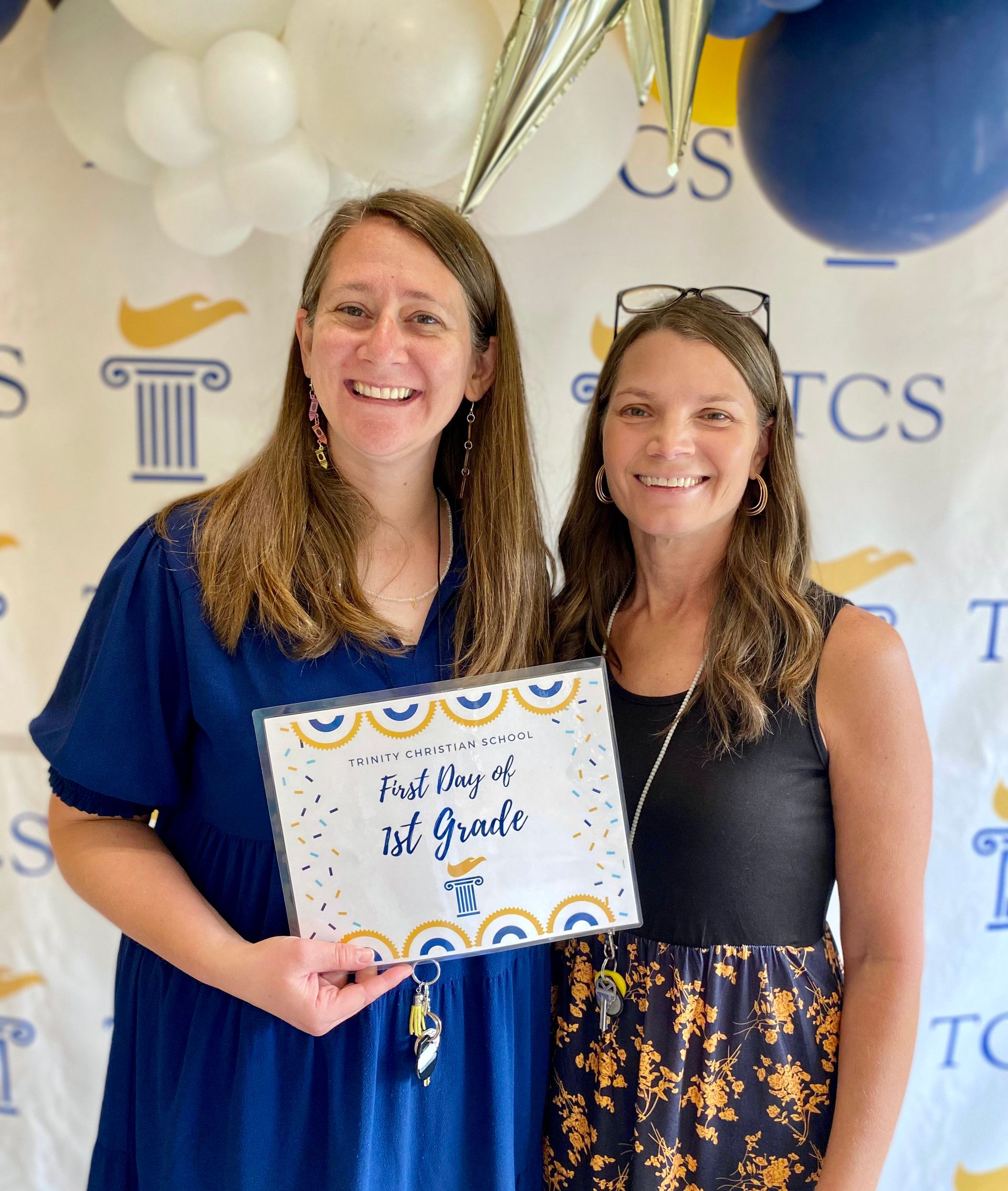 Two women smiling and holding a framed award at a blue-and-gold event backdrop with balloons