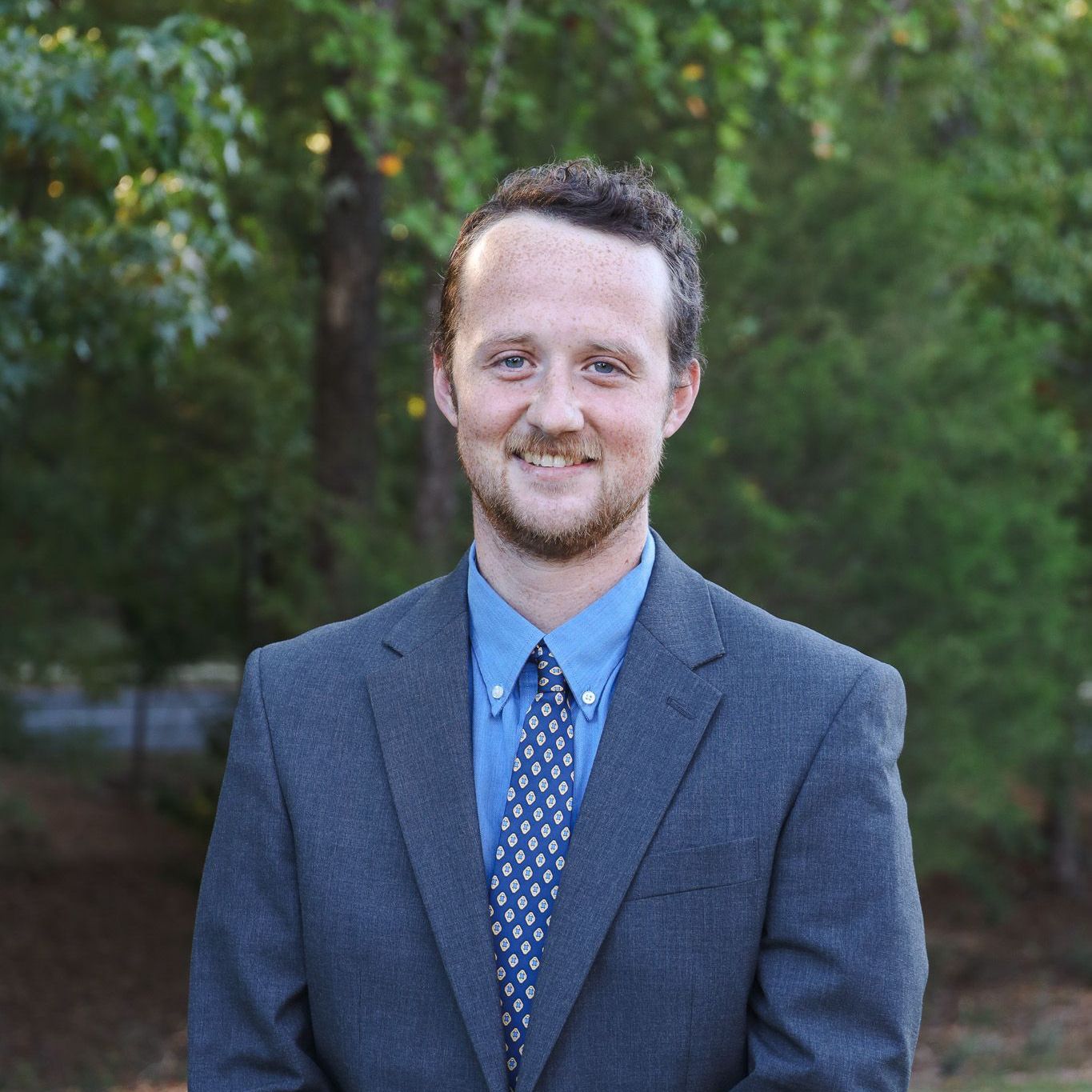 Portrait of a smiling man in a blue suit and tie standing outdoors with trees in the background