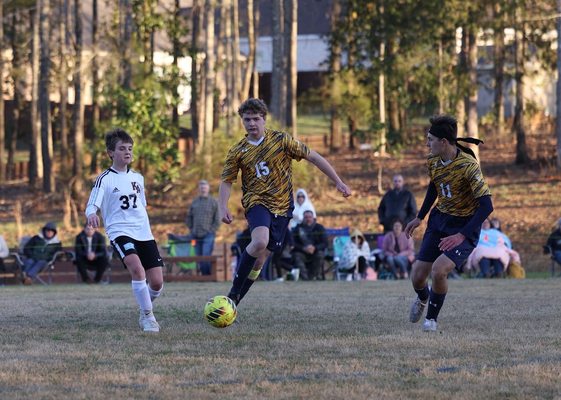 Three soccer players chase a yellow ball on a field with spectators in the background.